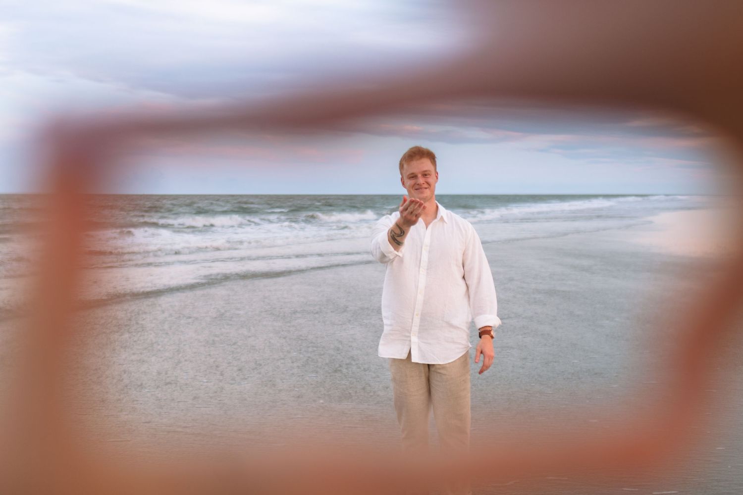 Smiling man in white linen shirt standing on beach framed by sunglasses.