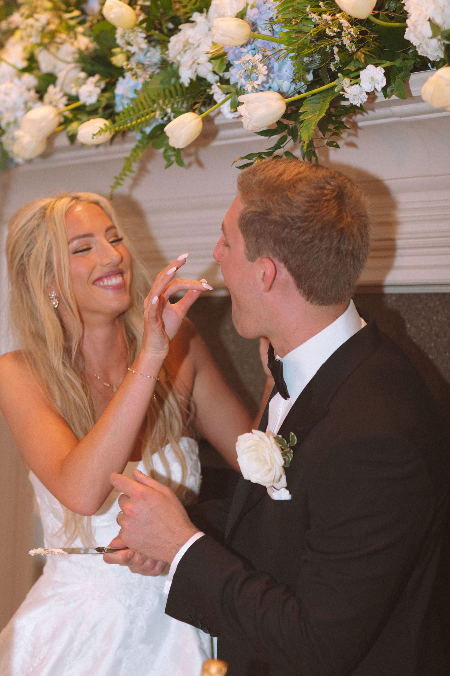 Blonde bride places wedding ring on laughing groom's finger beneath floral arrangement.
