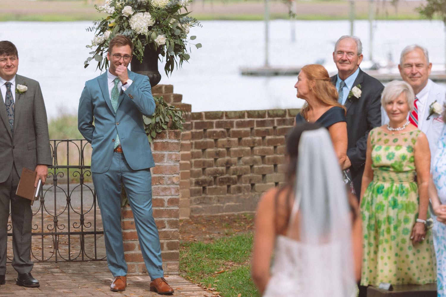 Emotional groom in teal suit reacts to seeing his bride walking down the aisle at outdoor waterfront wedding.
