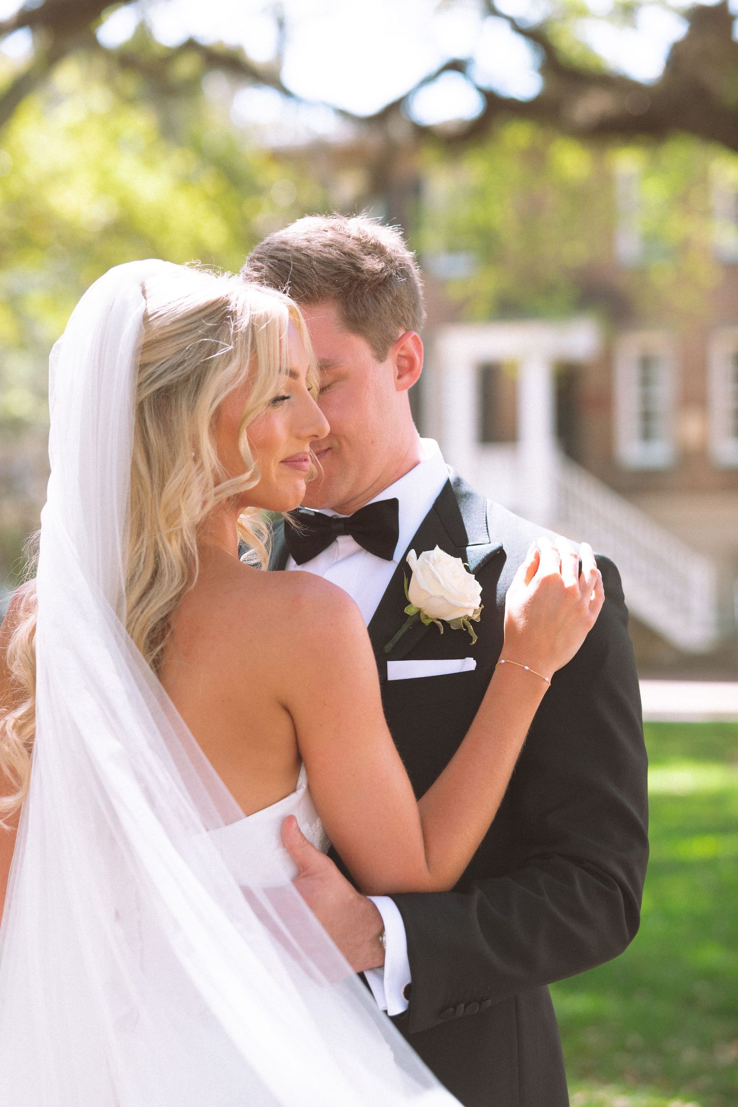 Bride and groom share a romantic embrace outdoors, with bride in white gown and veil, groom in classic black tuxedo.