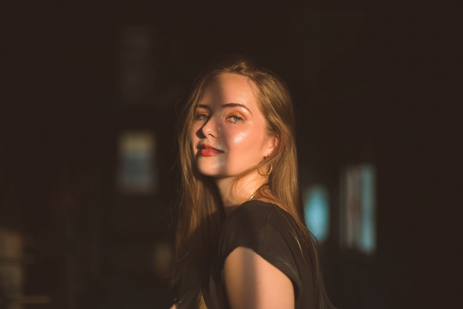 Young woman with long hair and a black top glances over her shoulder, her face lit by warm golden sunlight against a dark background.