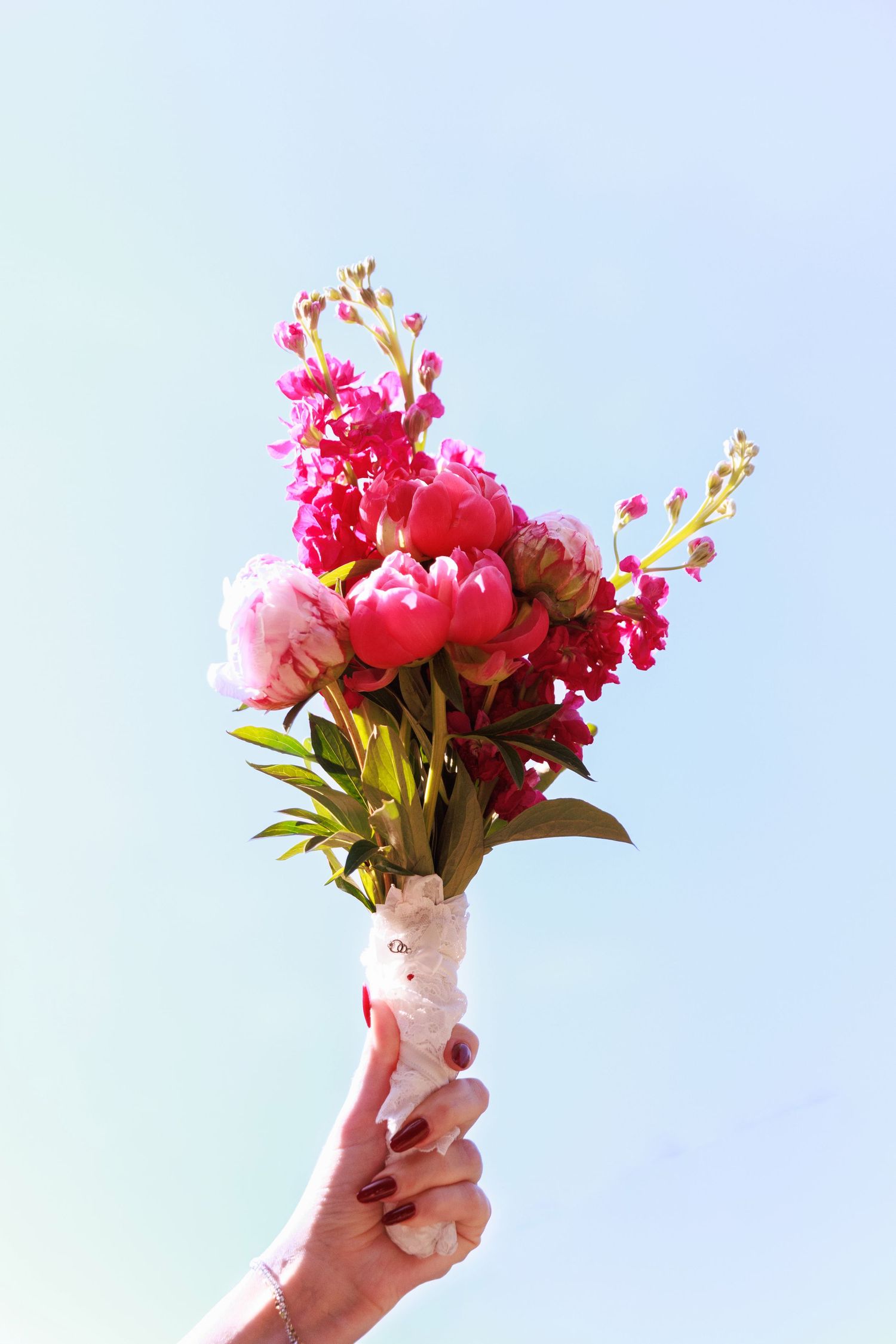 Hand holding a vibrant pink peony and snapdragon bridal bouquet against a clear light blue sky.