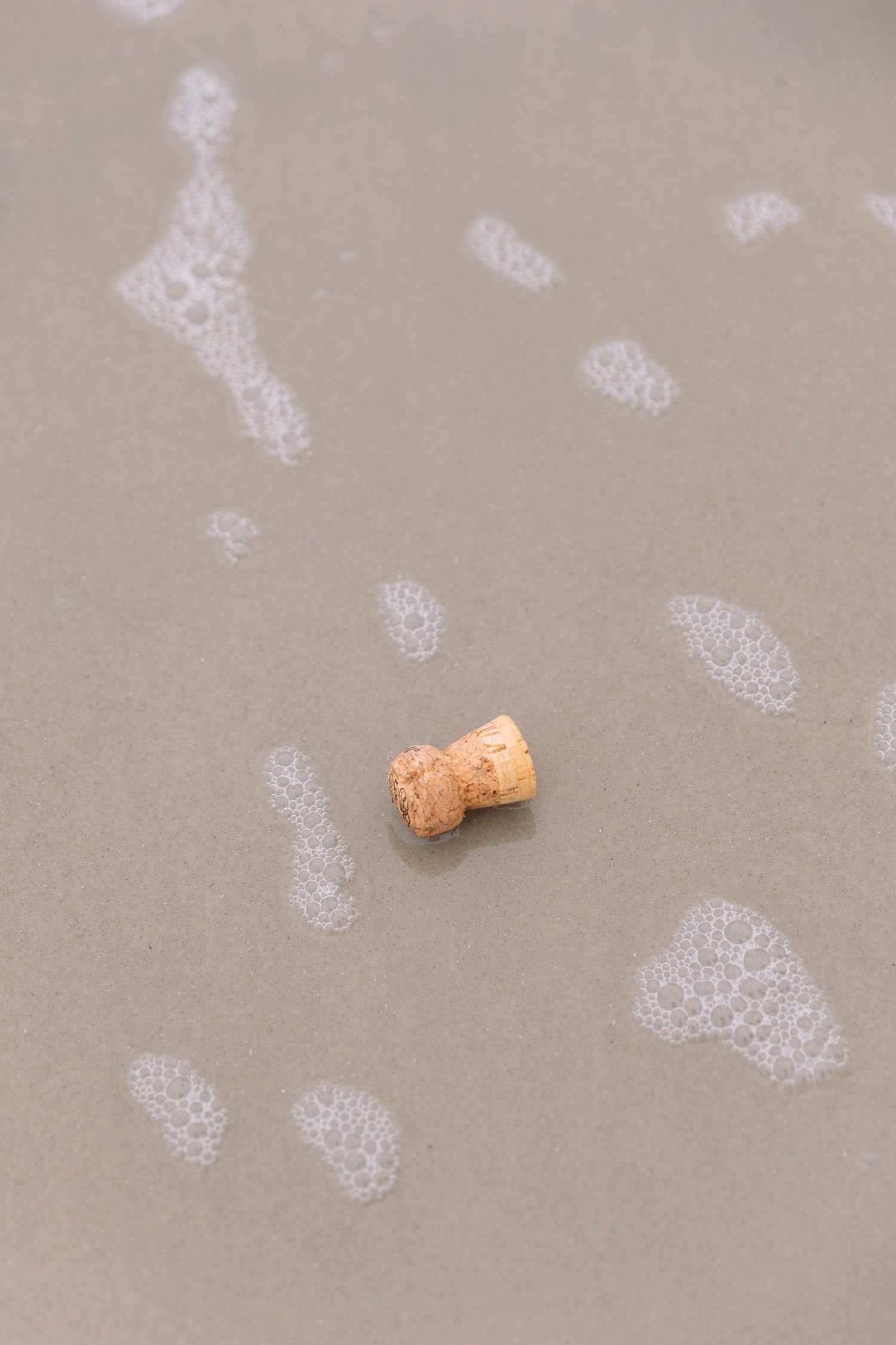 A champagne cork resting on wet sand surrounded by sea foam footprints at the beach.
