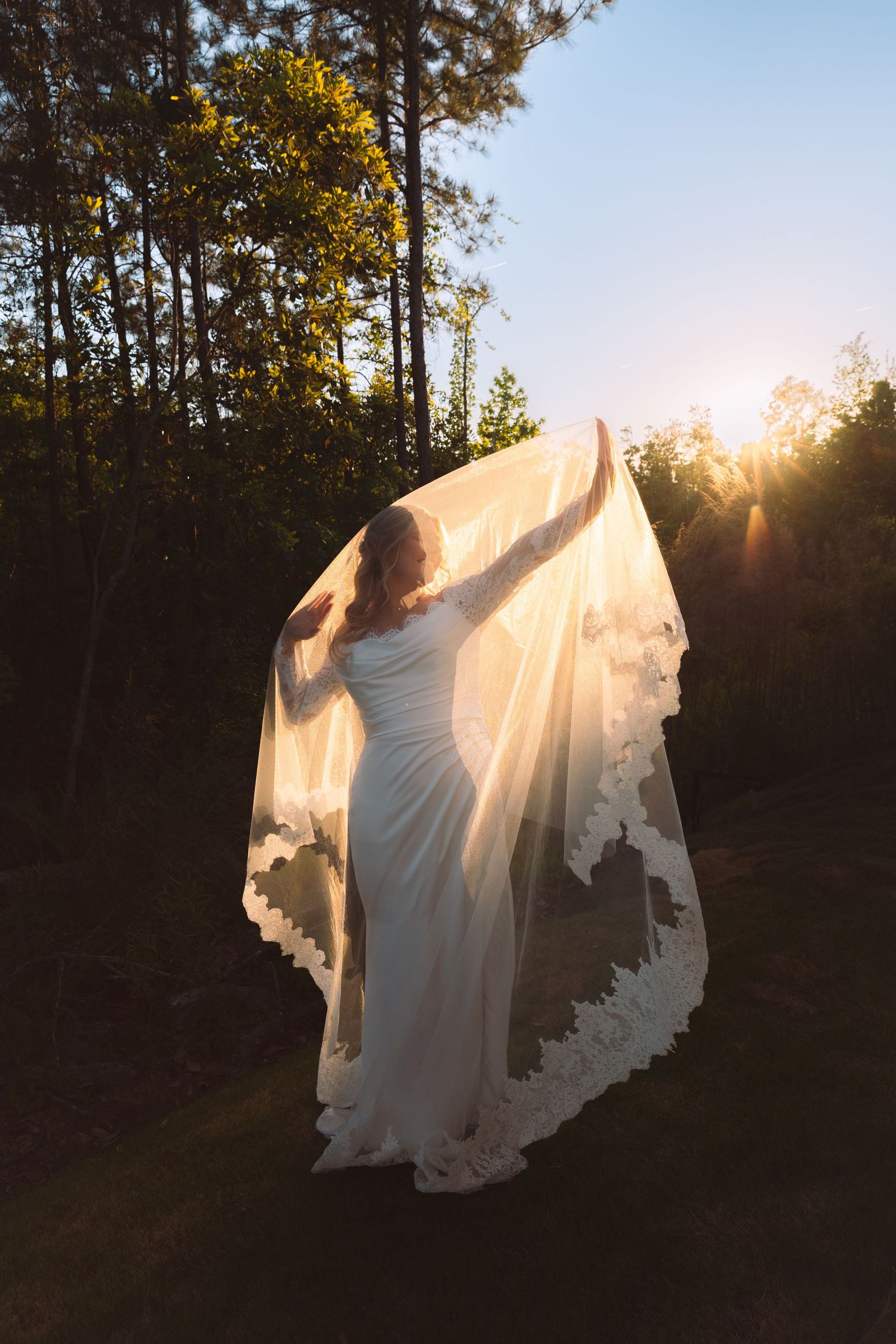 Bride in white gown and lace veil poses outdoors at golden sunset with trees in background.