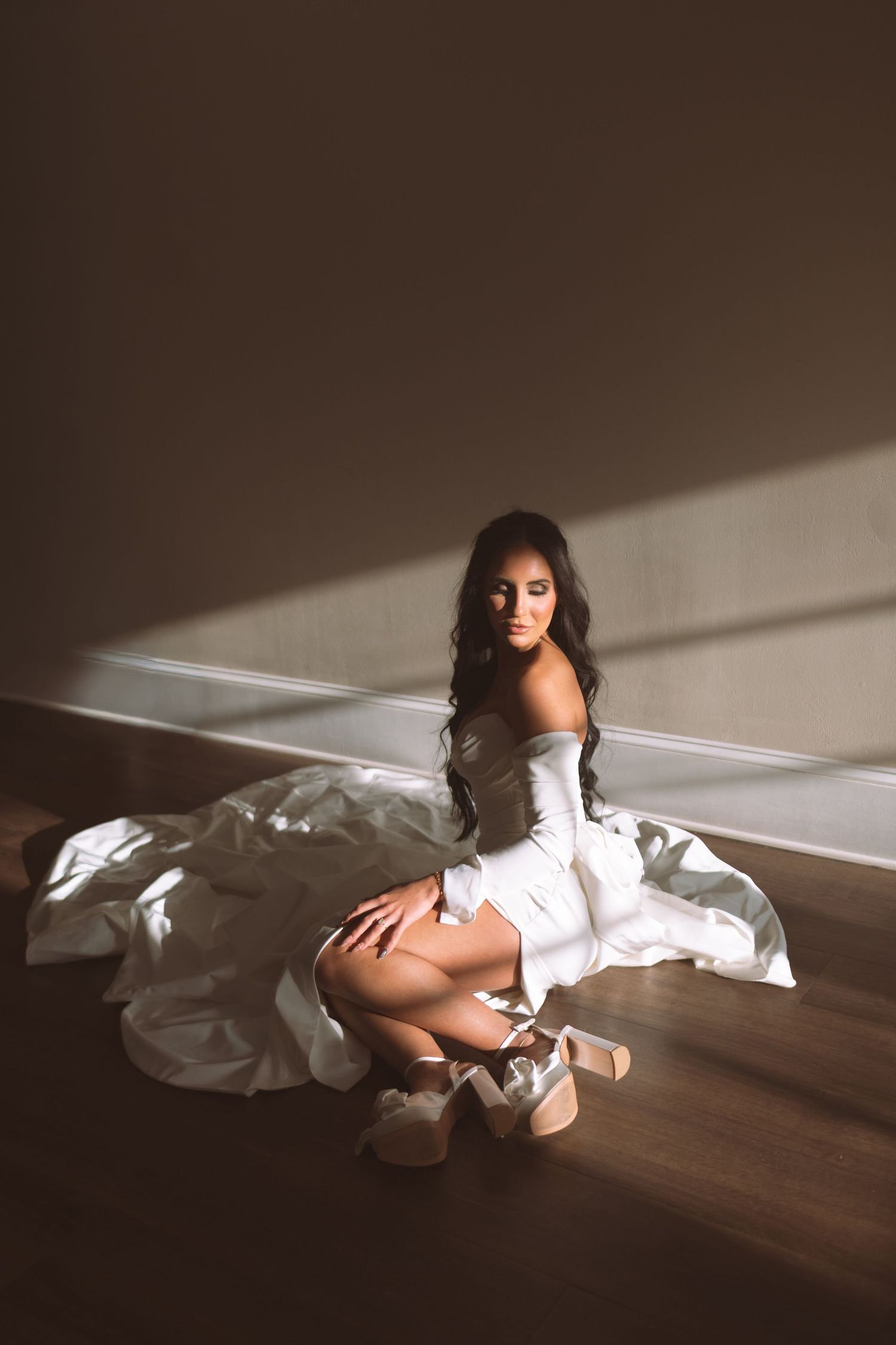Bride in white off-shoulder gown sitting gracefully on wooden floor in dramatic natural light.