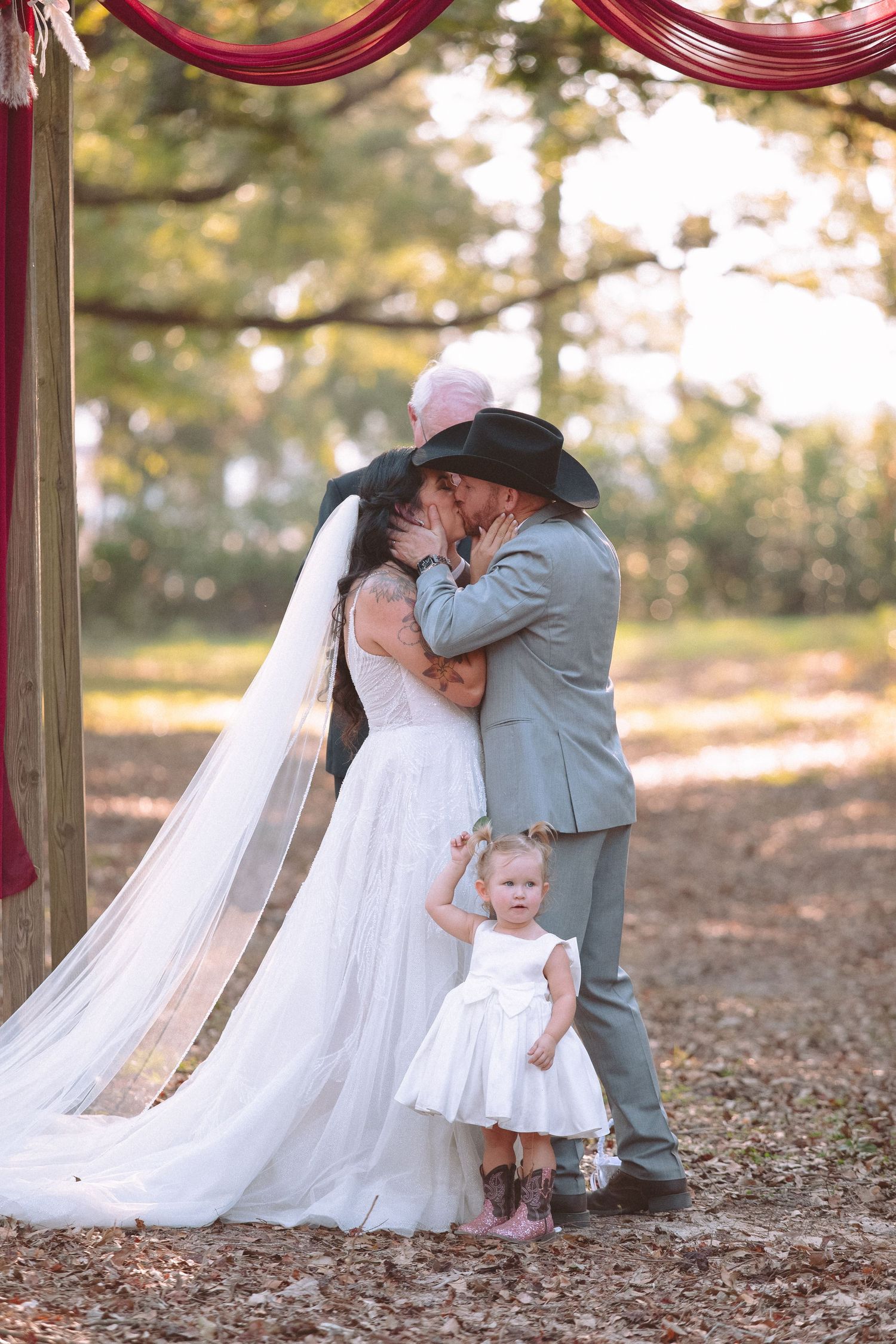 Bride and groom share first kiss with toddler flower girl at outdoor rustic wedding.