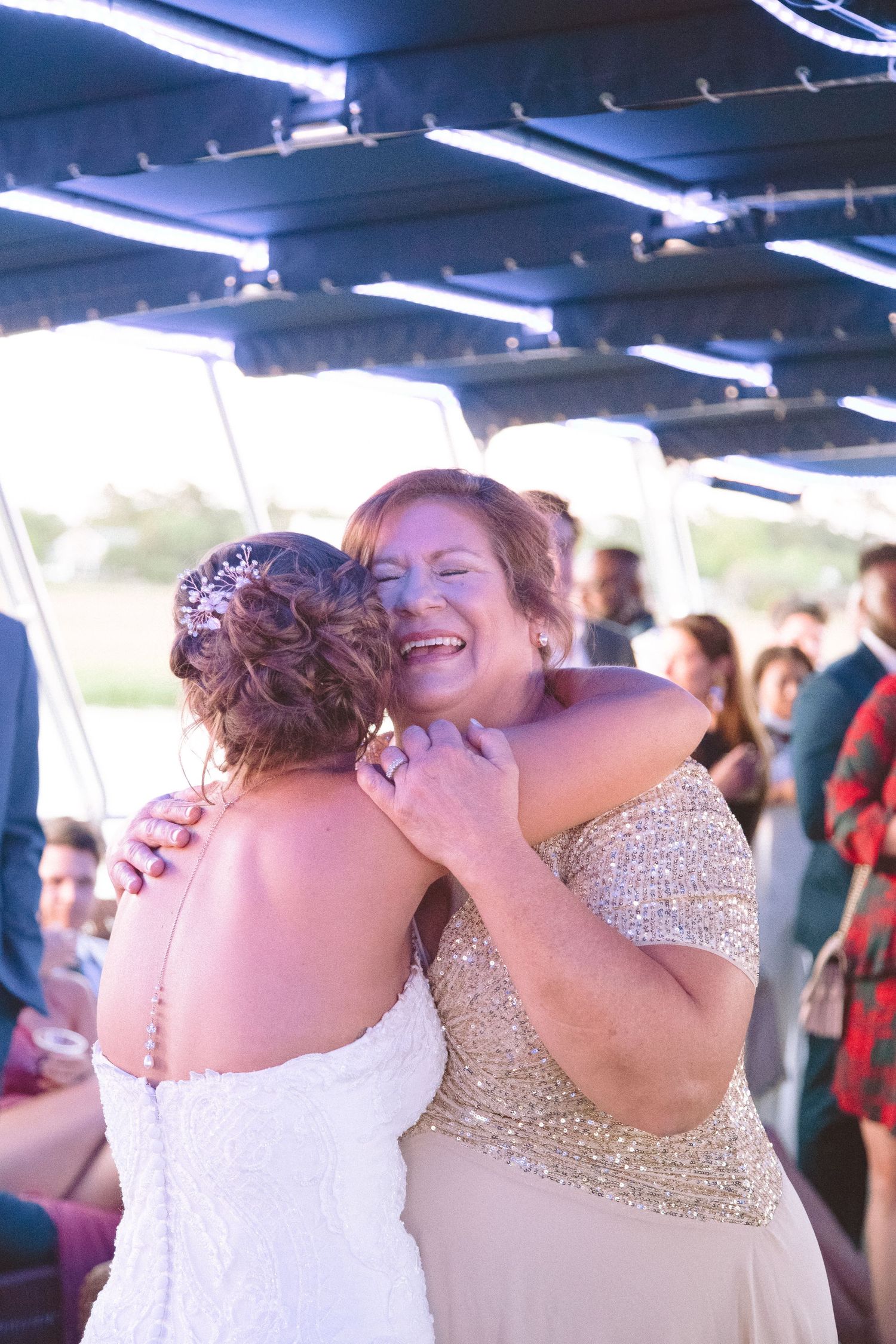 Joyful woman in gold sequin dress laughs while hugging bride at wedding reception.