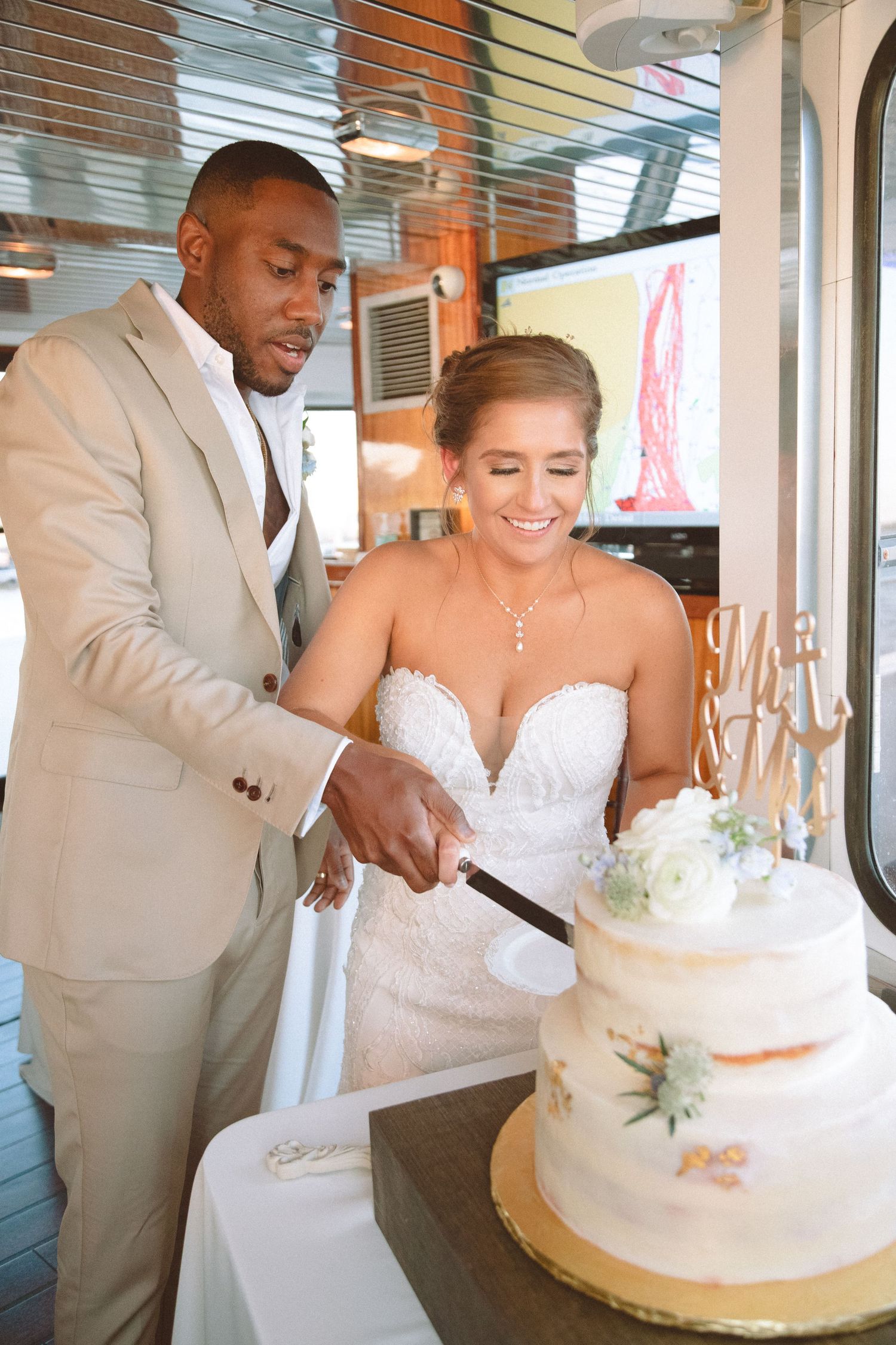 Bride laughing joyfully as couple cuts white two-tier wedding cake at nautical boat reception.