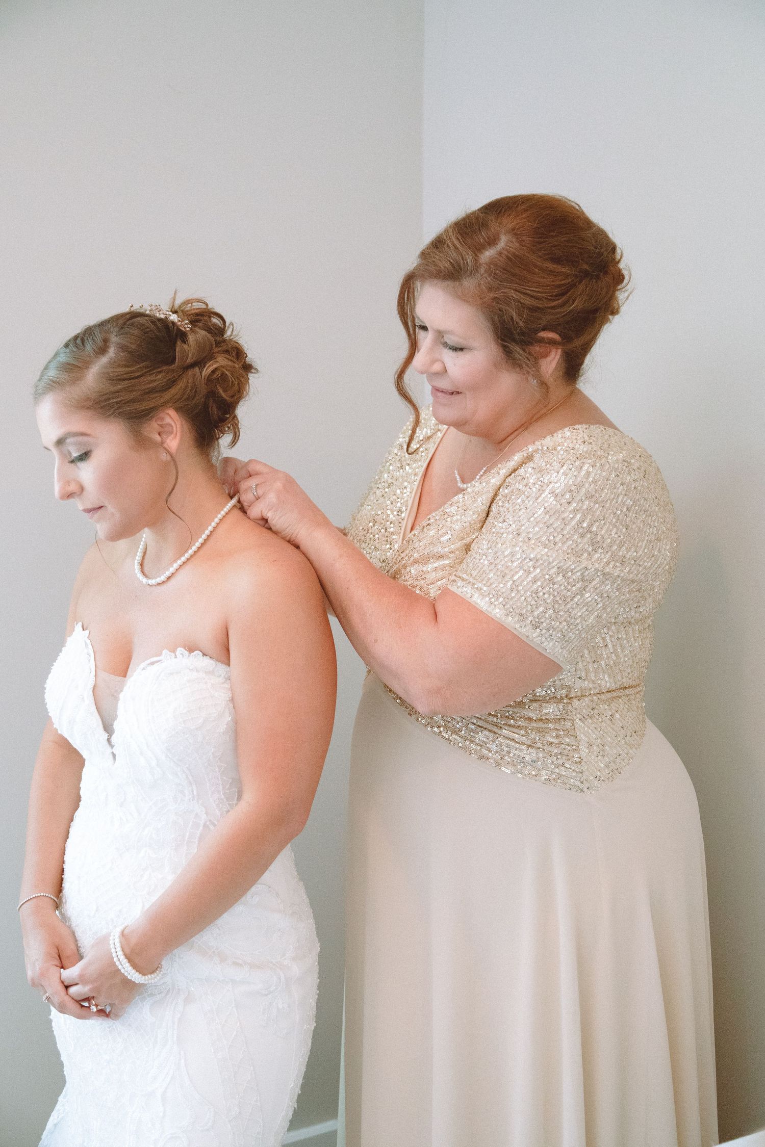 Mother fastening pearl necklace on bride wearing strapless lace wedding gown before ceremony.