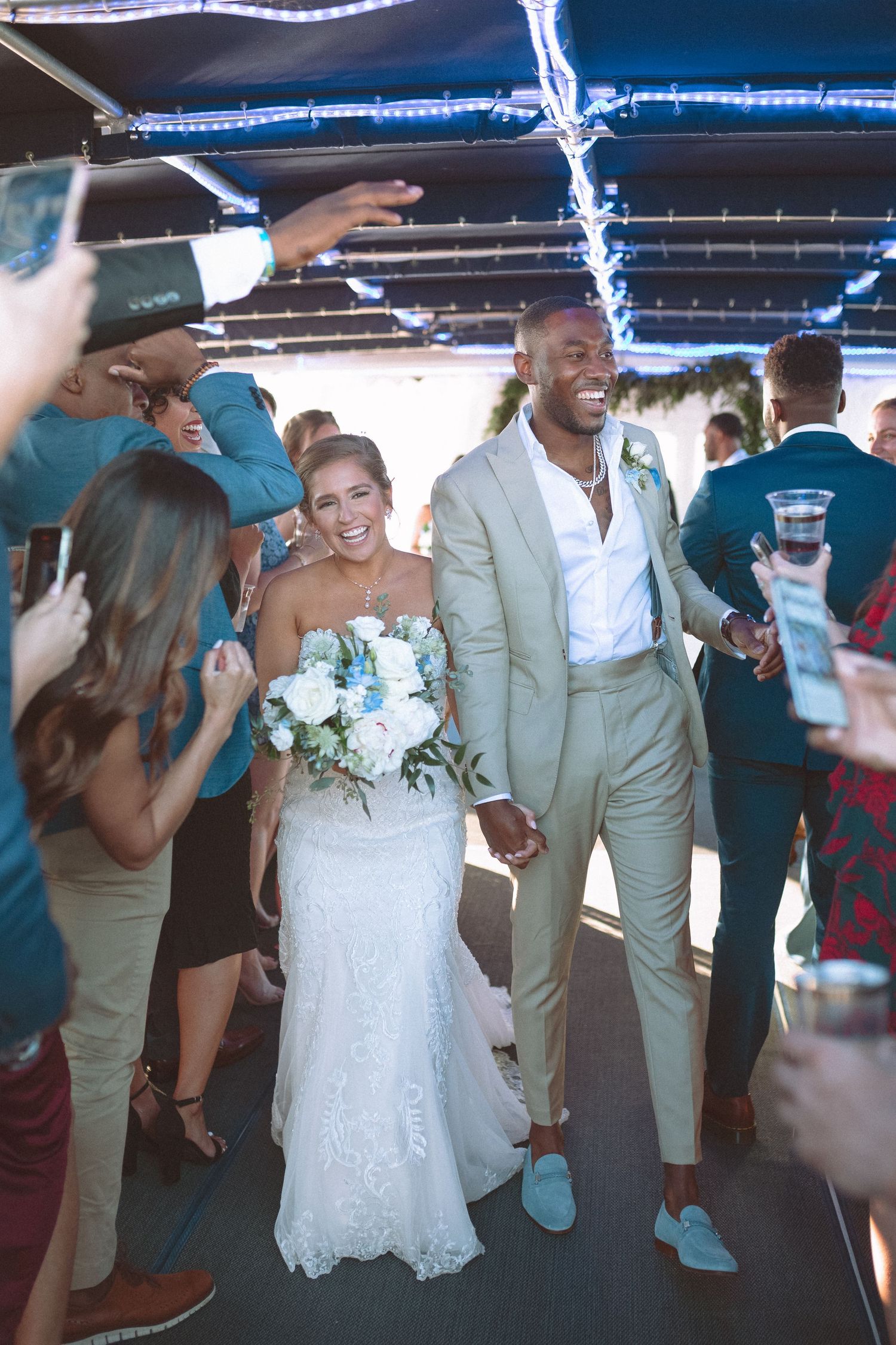 Joyful newlyweds walk through cheering guests at their wedding reception, bride holding white bouquet.