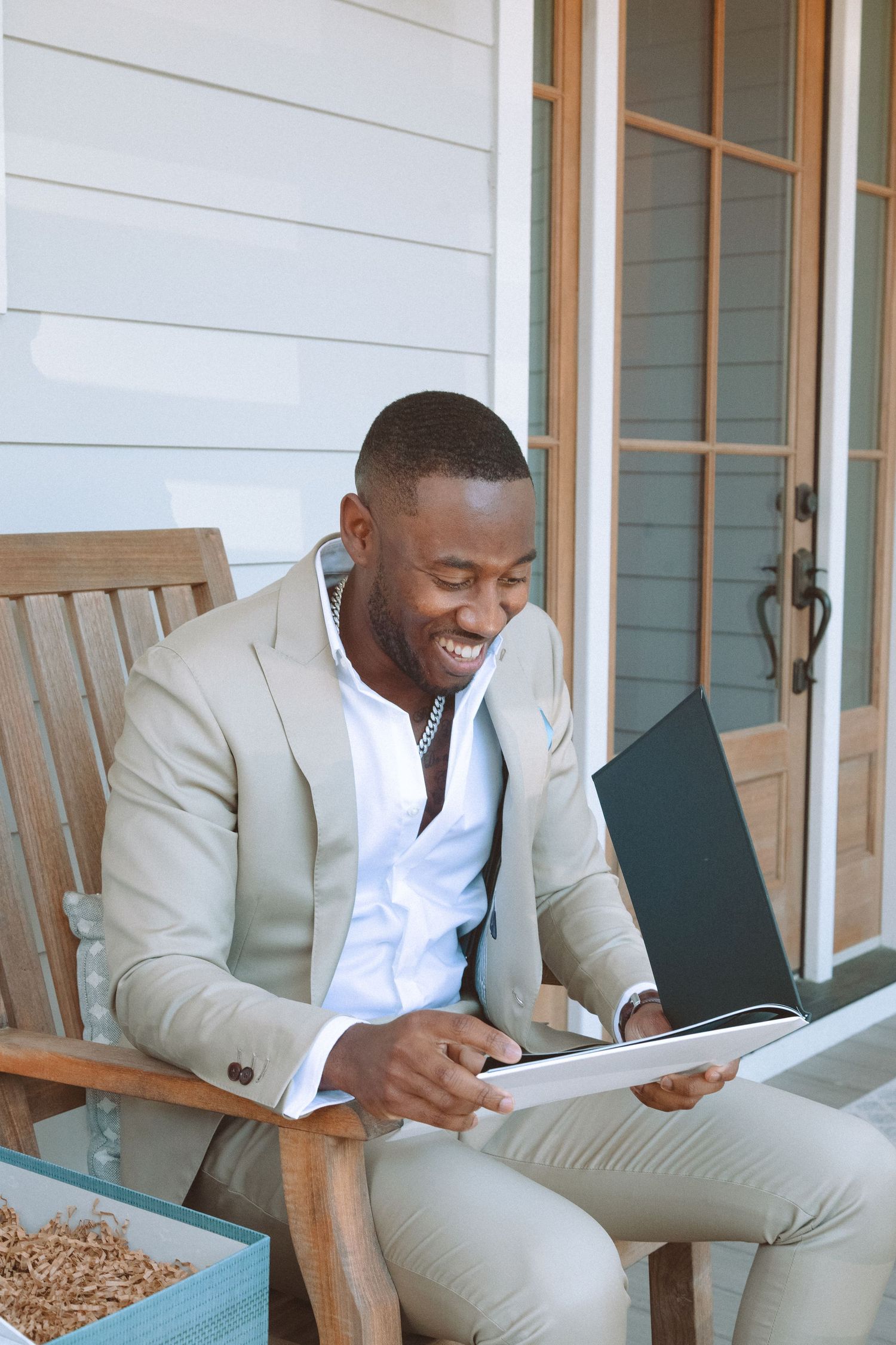 Joyful man in tan suit laughing while reviewing a black album seated in wooden chair on porch.