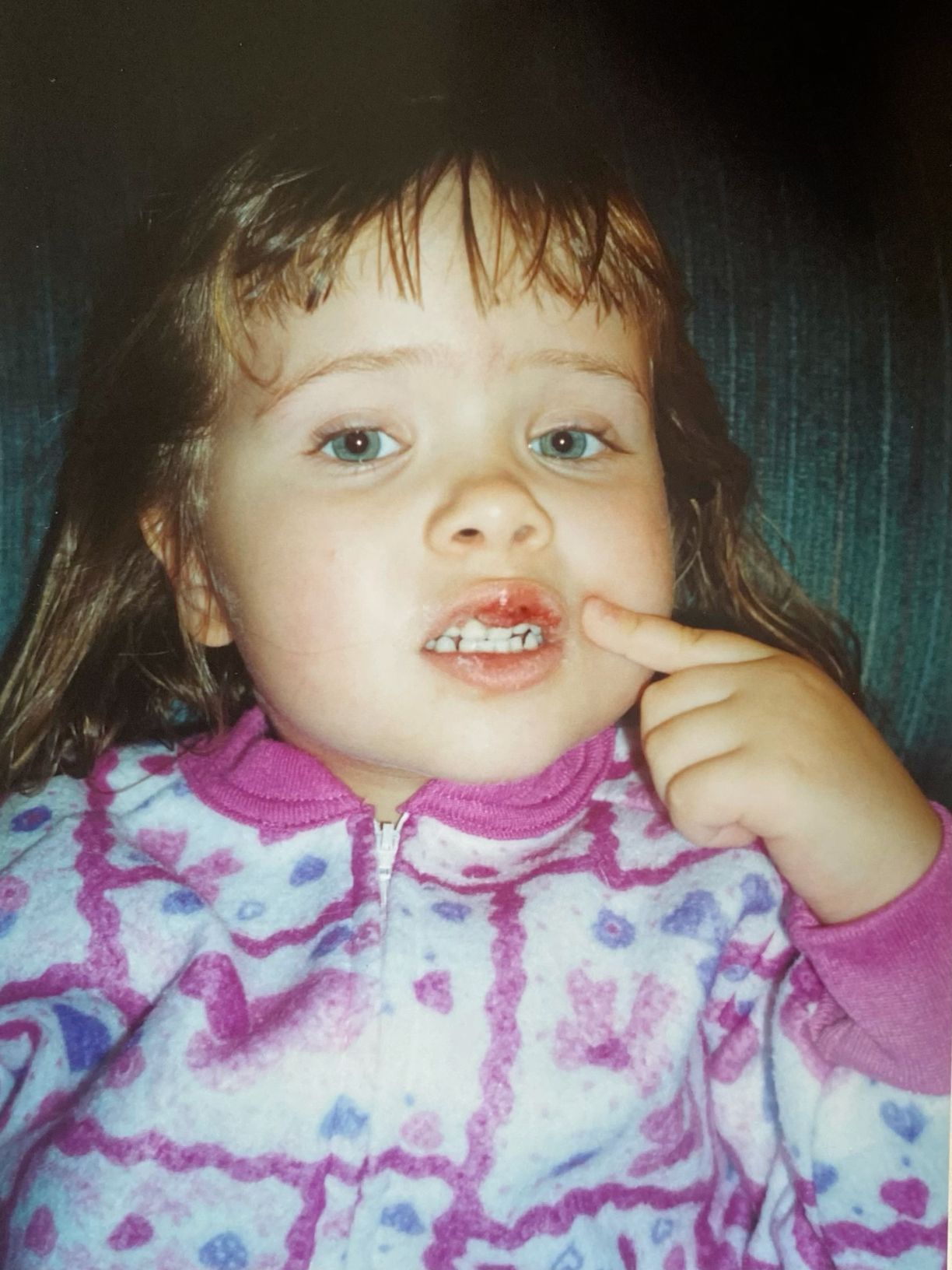 A close-up portrait of a young toddler girl with brown hair pointing at her mouth.