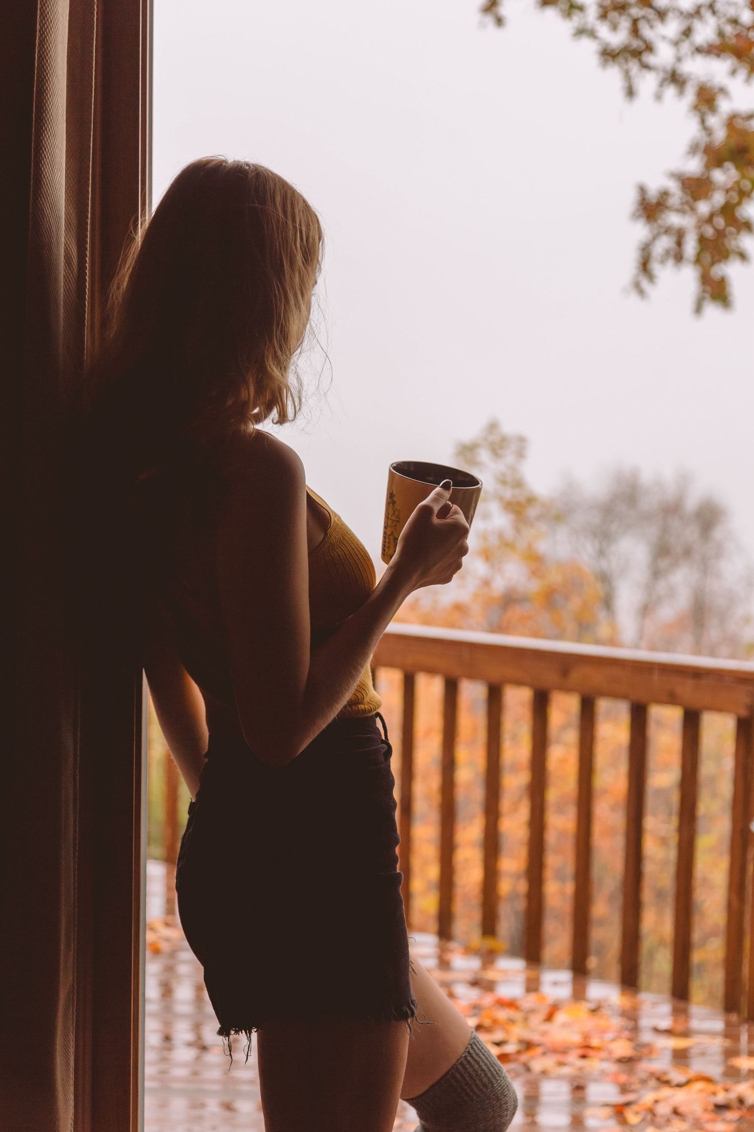 Woman silhouetted in doorway holding a mug on a rainy autumn day.