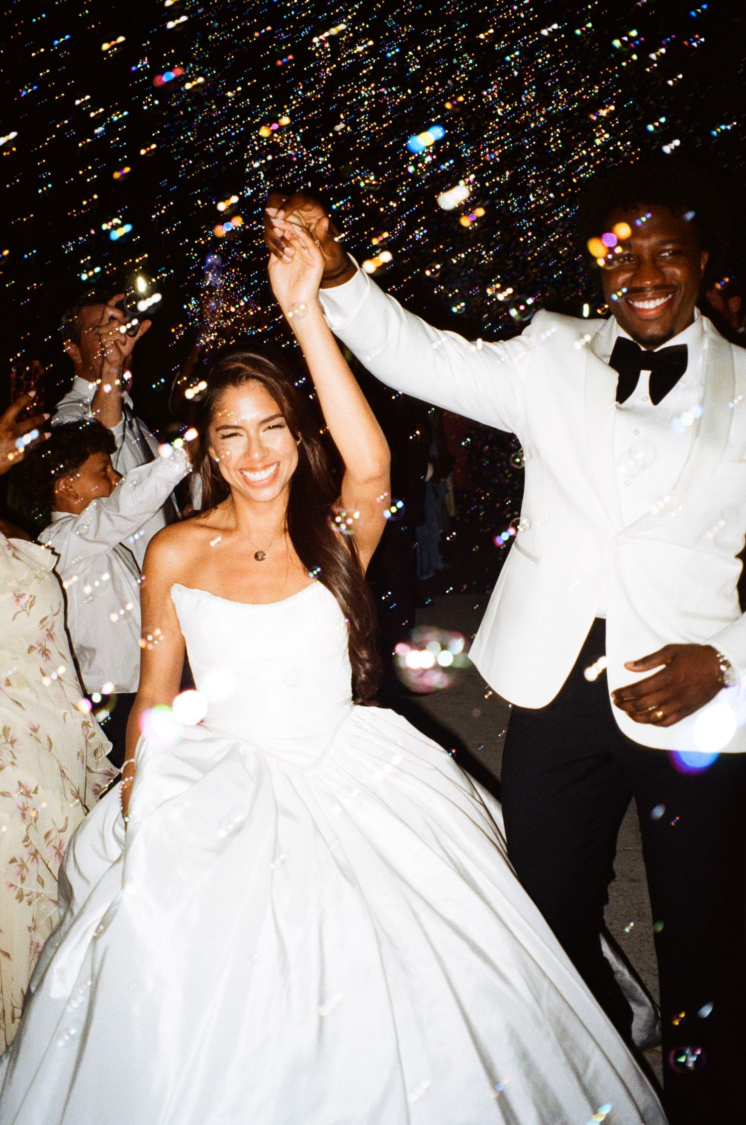 Joyful newlyweds celebrate at night wedding reception, bride in white gown and groom in white tuxedo with bow tie.