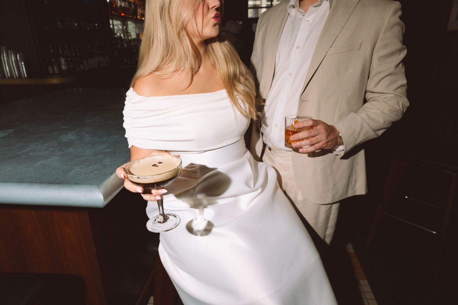 Bride in white off-shoulder dress holding cocktail beside groom in beige suit at bar.