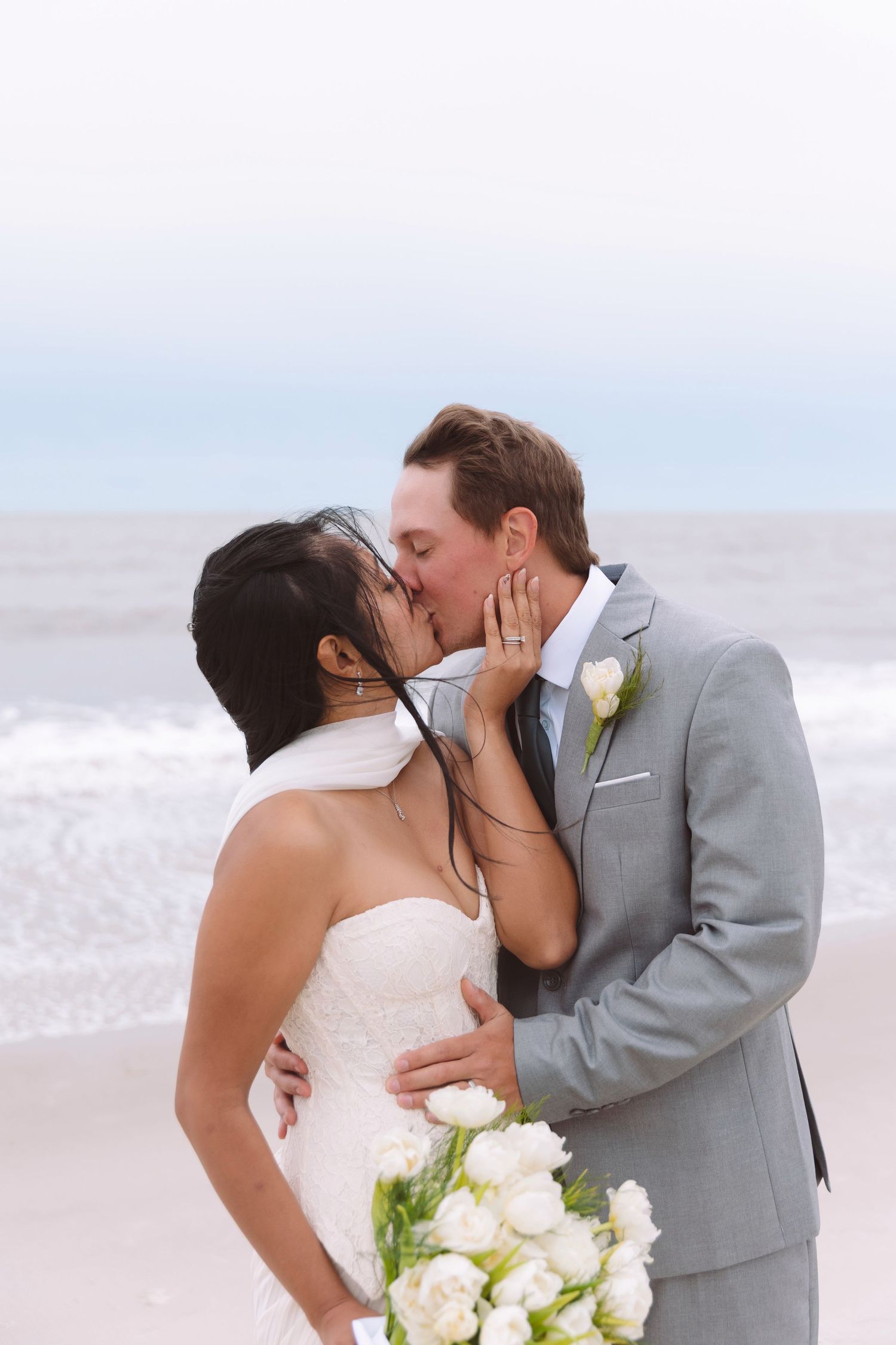 Bride and groom share a kiss on the beach, she holds white tulip bouquet, he wears gray suit.