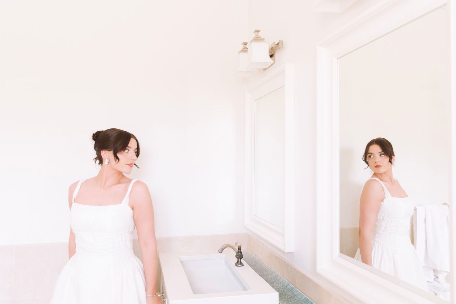 Elegant bride in a white gown admiring her reflection in a large mirror in a bright bathroom.