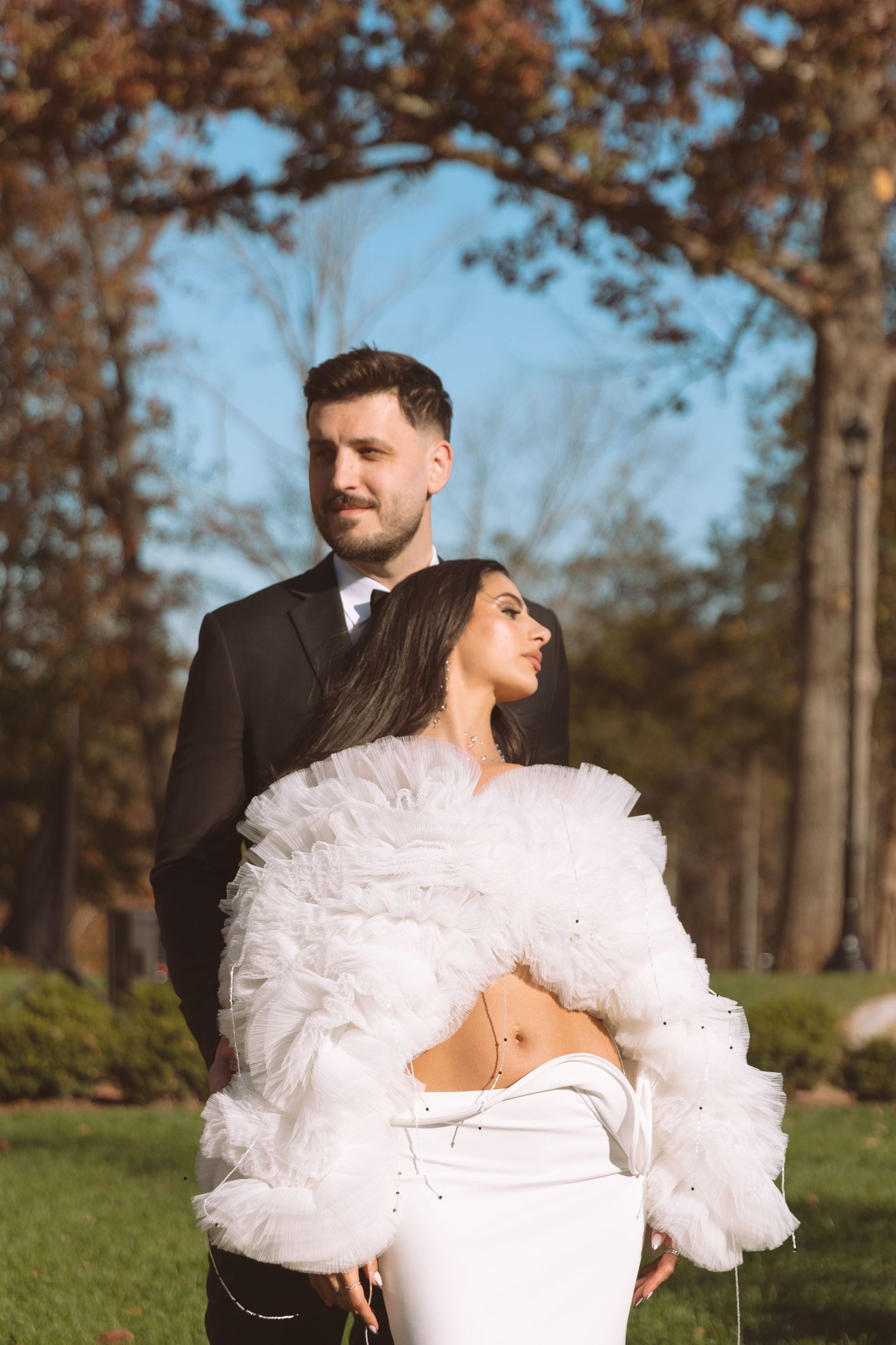 Couple poses outdoors in autumn park, bride wearing ruffled white jacket and groom in black suit.