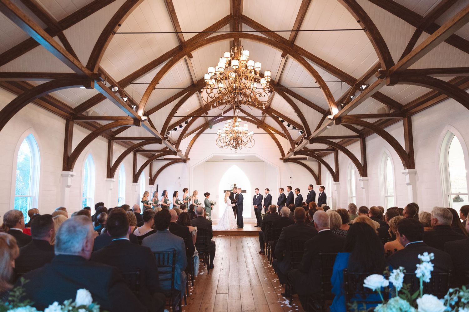 Beautiful wedding ceremony inside a white chapel with vaulted wooden beams and chandelier lighting.