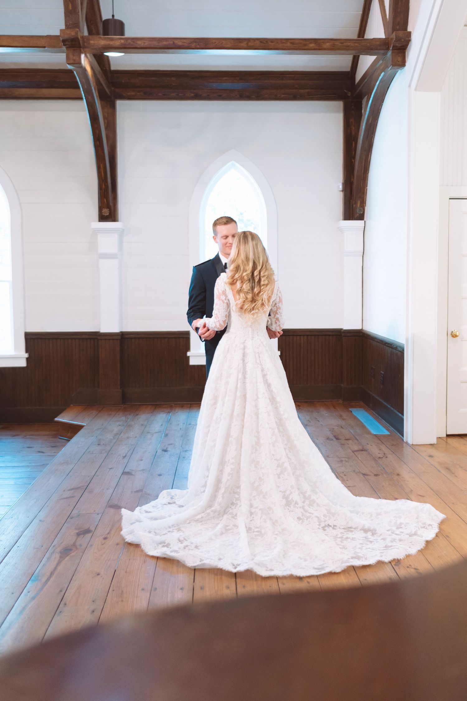 Bride and groom stand in sunlit white chapel with wooden beams and cathedral windows.