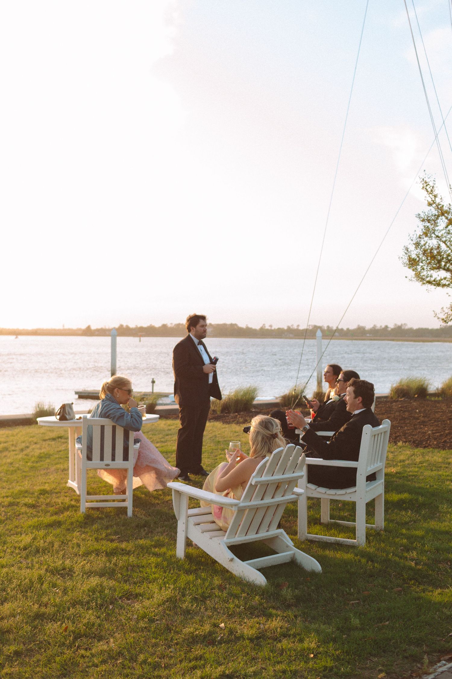 Wedding guests relaxing in white Adirondack chairs by the waterfront at sunset.