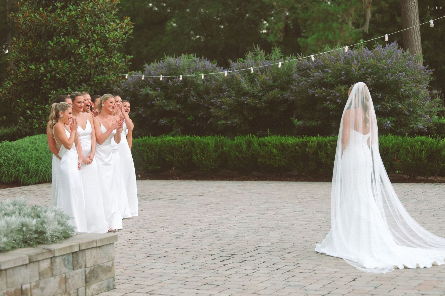 Bride in white gown and veil faces bridesmaids in white dresses on a brick patio surrounded by lush greenery.