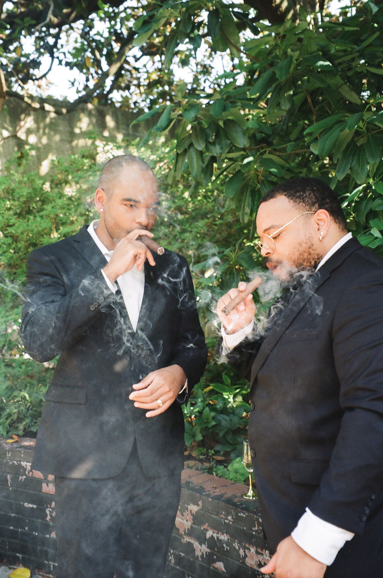 Two men in suits smoking cigars outdoors, surrounded by lush green trees, with smoke swirling around them.