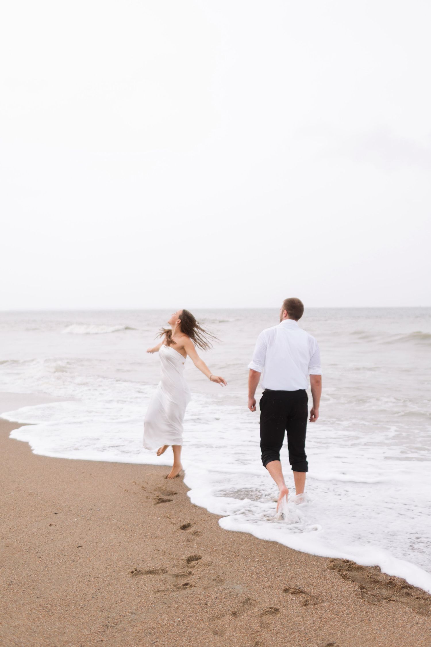 Couple walking barefoot along sandy beach shoreline, woman in white dress and man in white shirt with black pants.