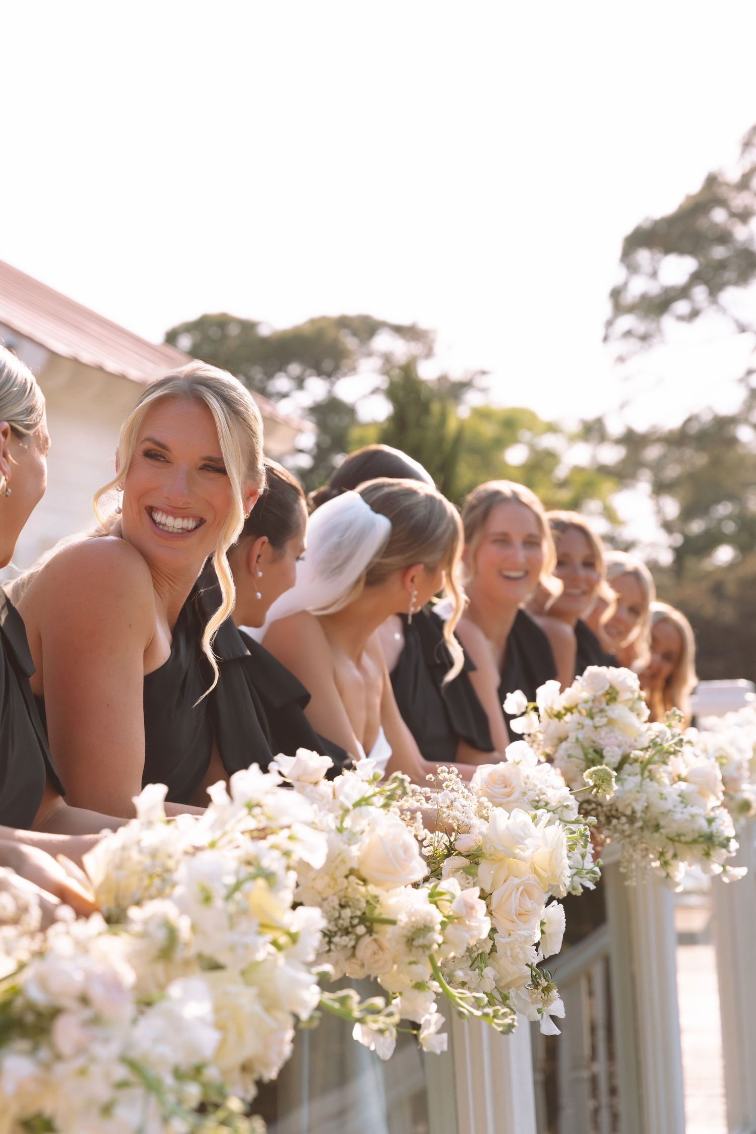 Smiling bridesmaids in black dresses holding white floral bouquets line a railing at an outdoor wedding ceremony.