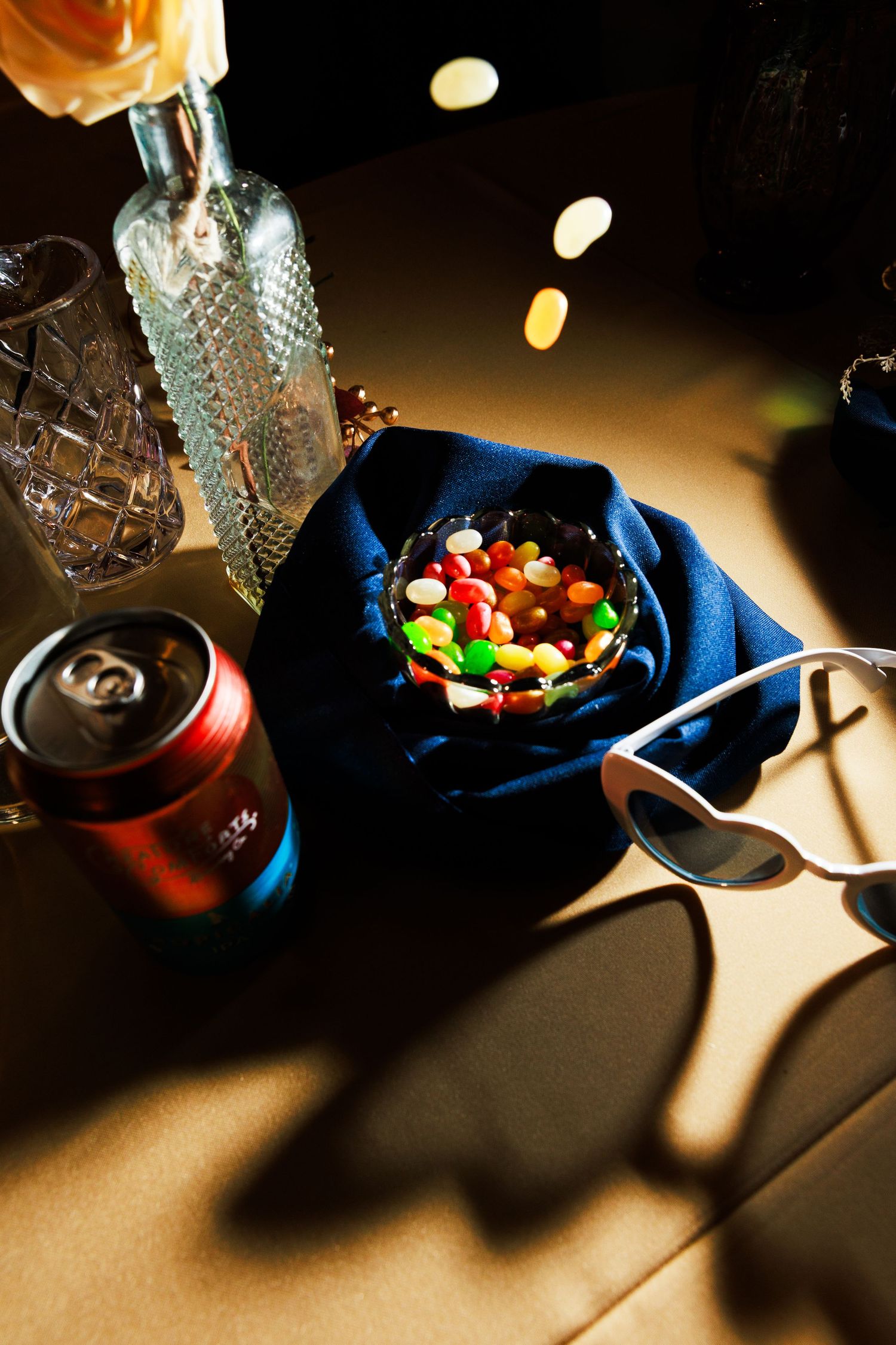 Colorful Skittles candy in a dark bowl on blue fabric beside sunglasses and a red soda can on a table.