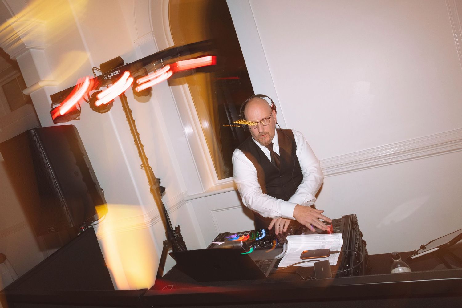 A bald DJ wearing glasses and a vest works at his equipment setup under warm red lighting at an indoor event.