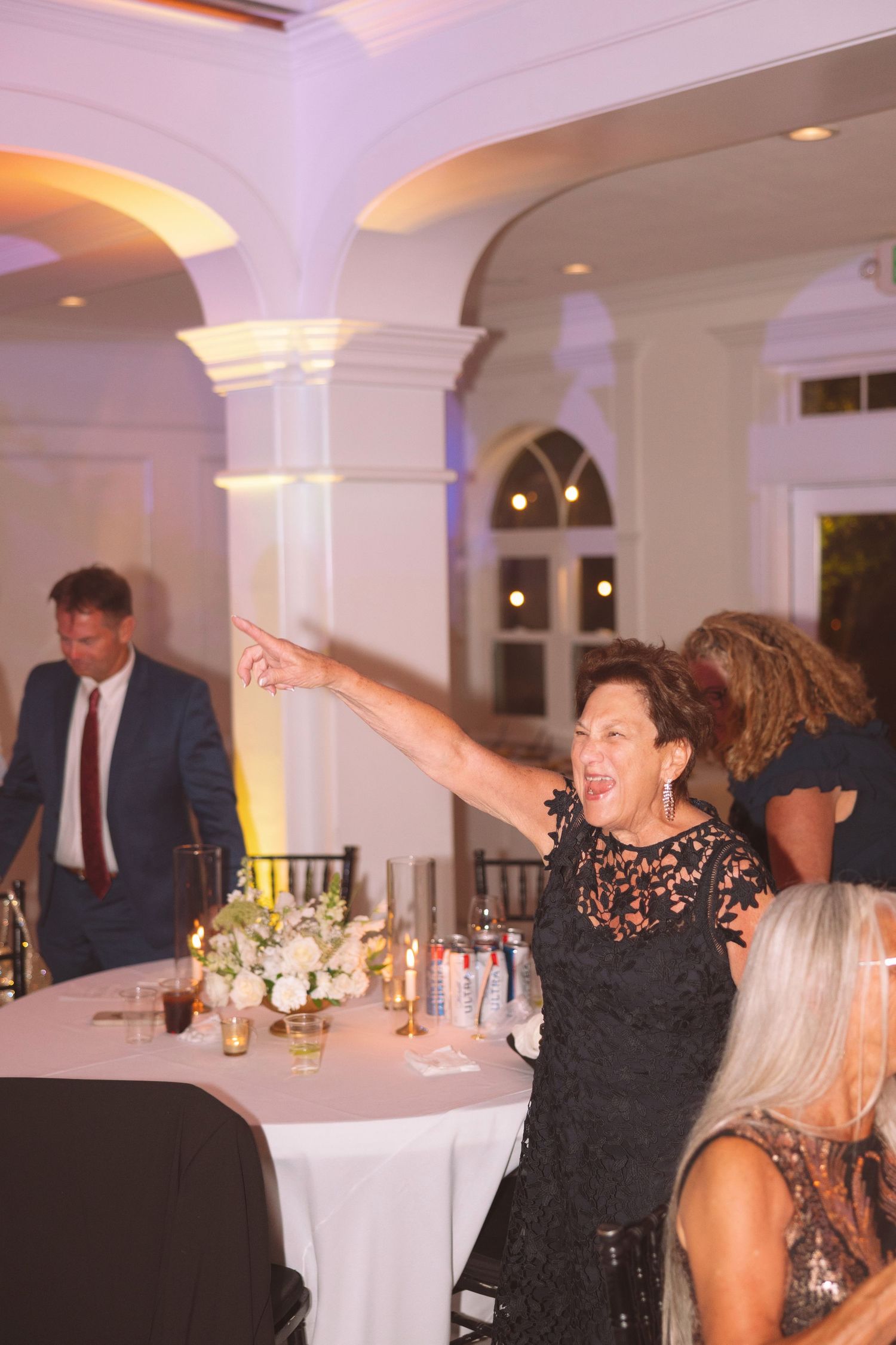 Woman in black lace dress pointing and laughing at elegant wedding reception with white columns.
