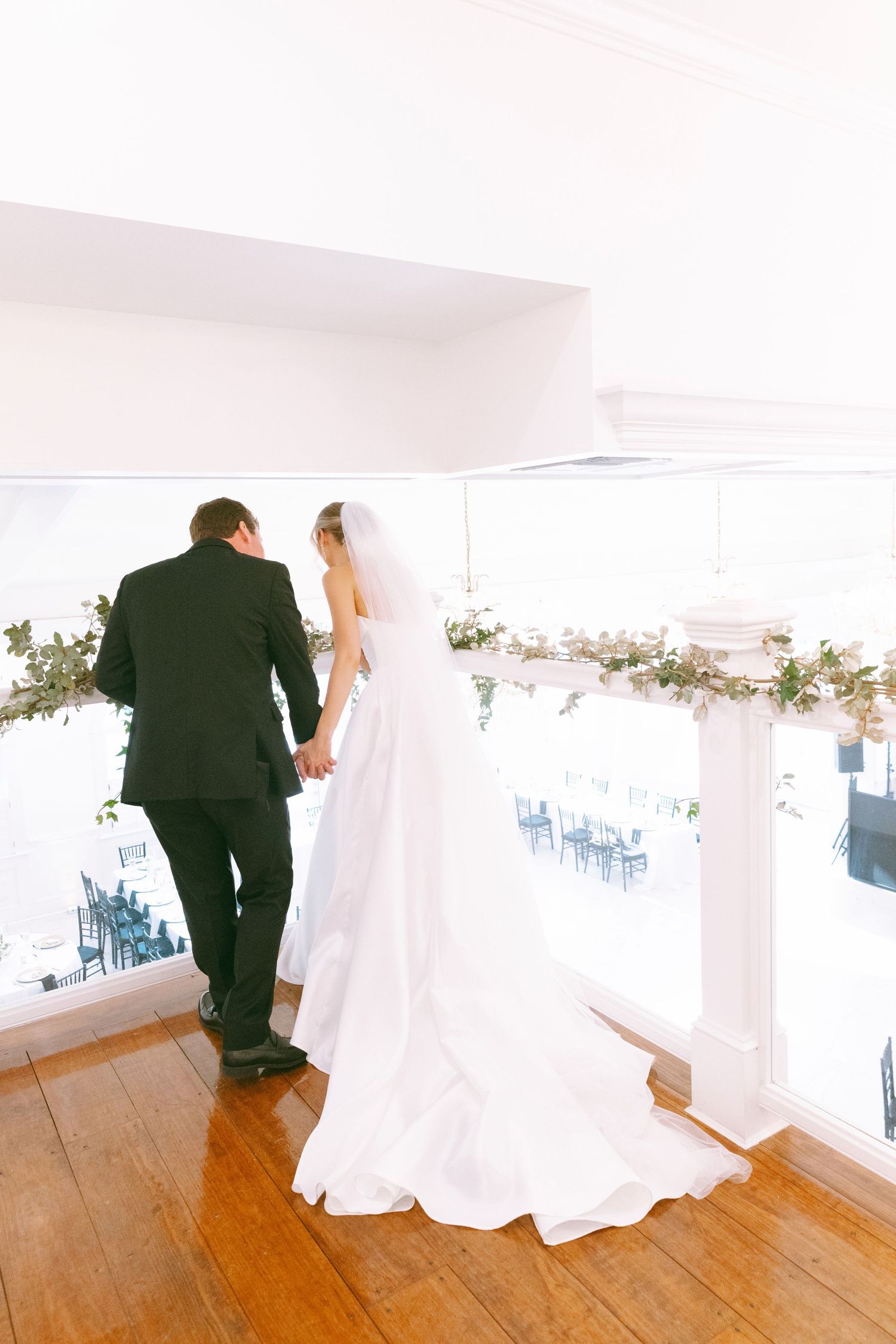 Bride and groom holding hands on a balcony overlooking a wedding reception venue, adorned with floral garlands.