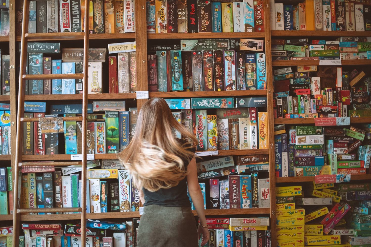 Young woman with long hair browsing colorful board games on wooden shelves in a game store.