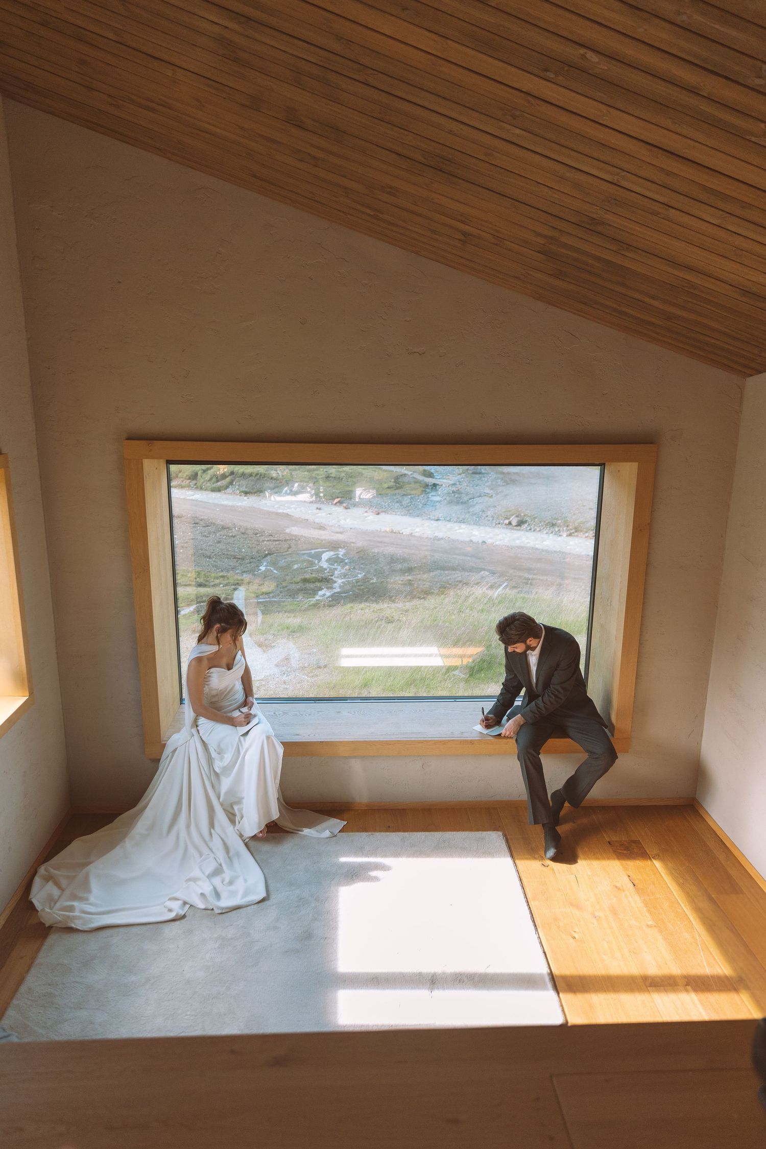 Bride and groom sitting by a large window with scenic mountain views on their wedding day.