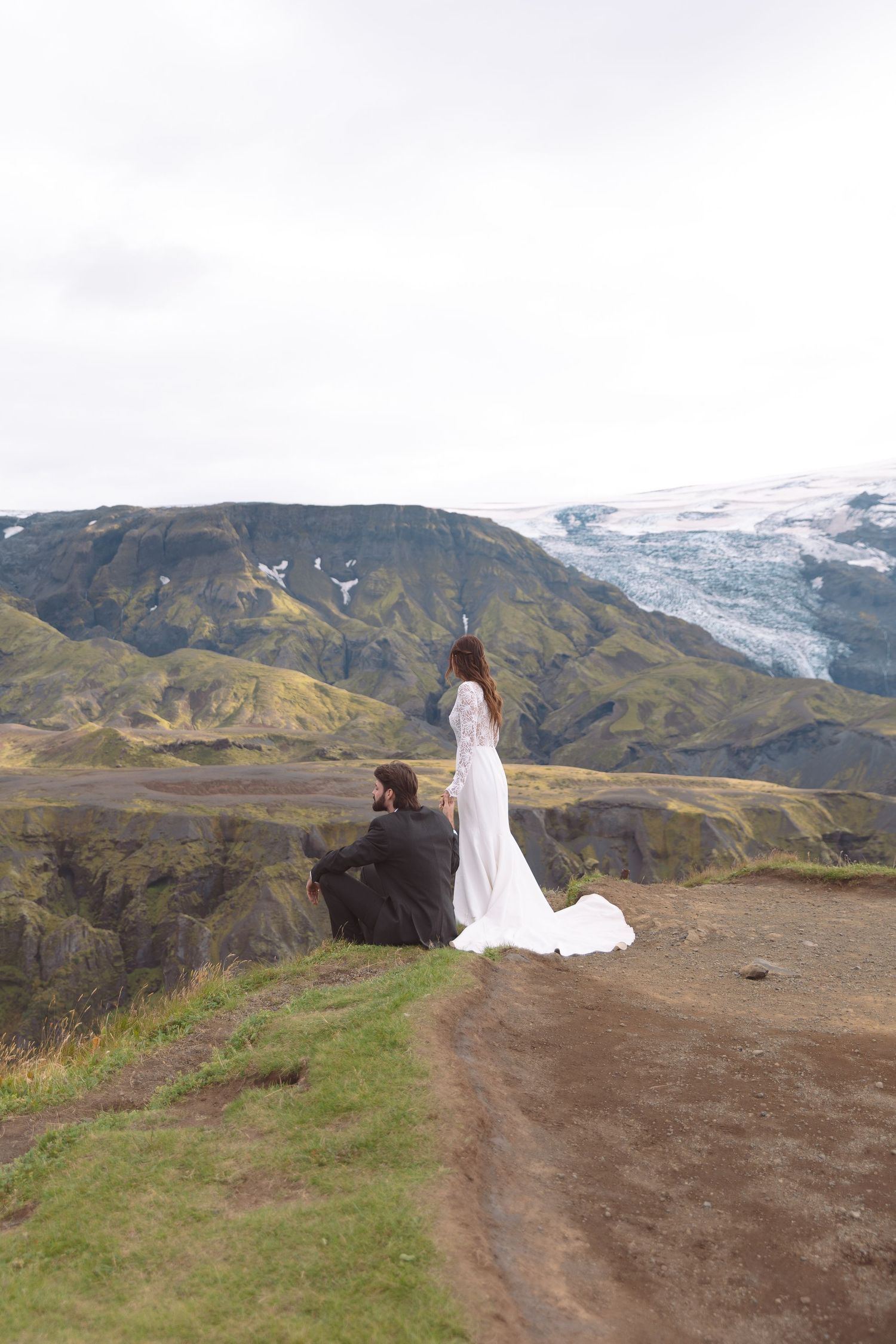 Bride in white gown and groom sit on Iceland cliff edge overlooking a stunning glacier landscape.