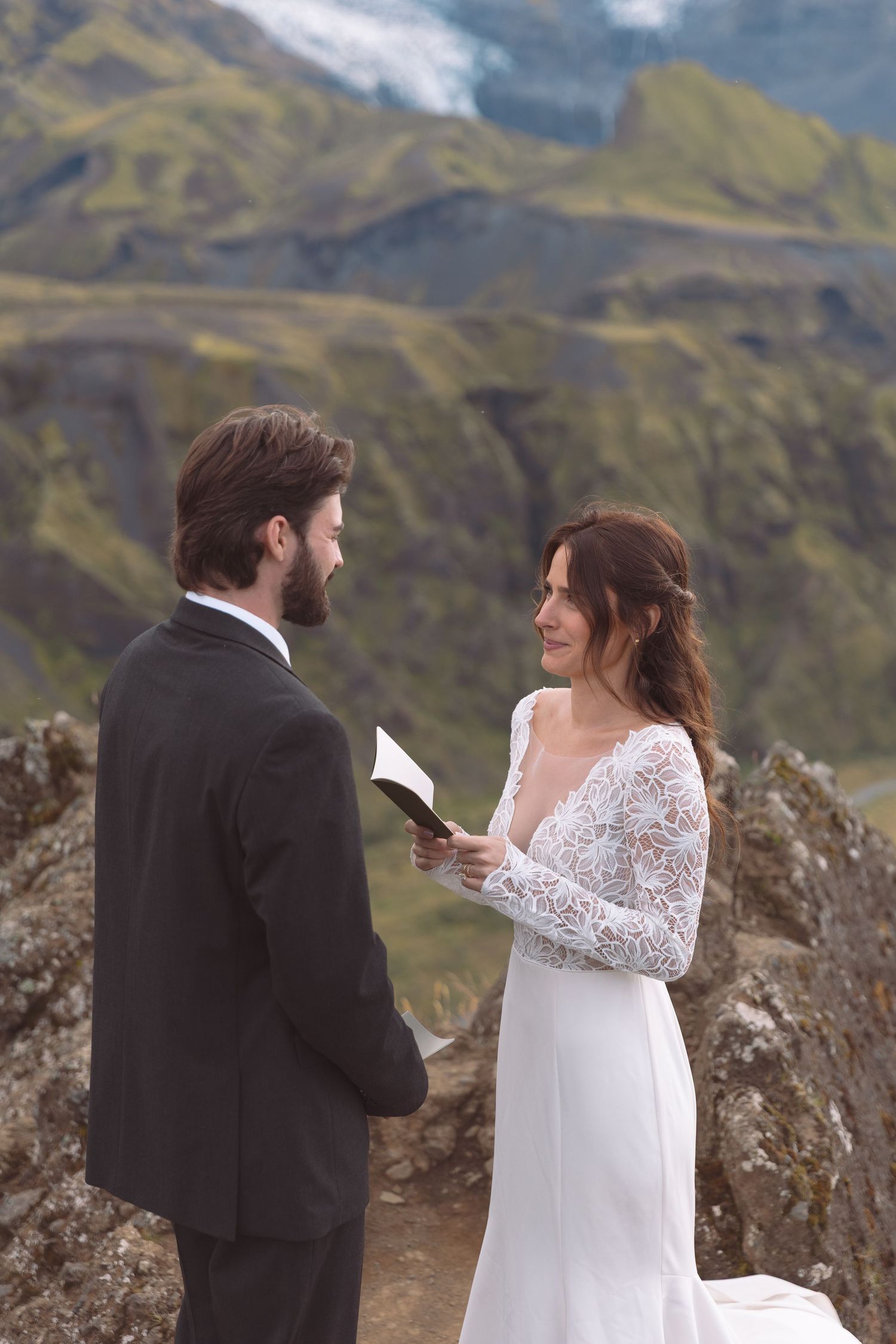Bride in lace wedding gown smiles while reading vows to bearded groom on scenic Icelandic mountainside.