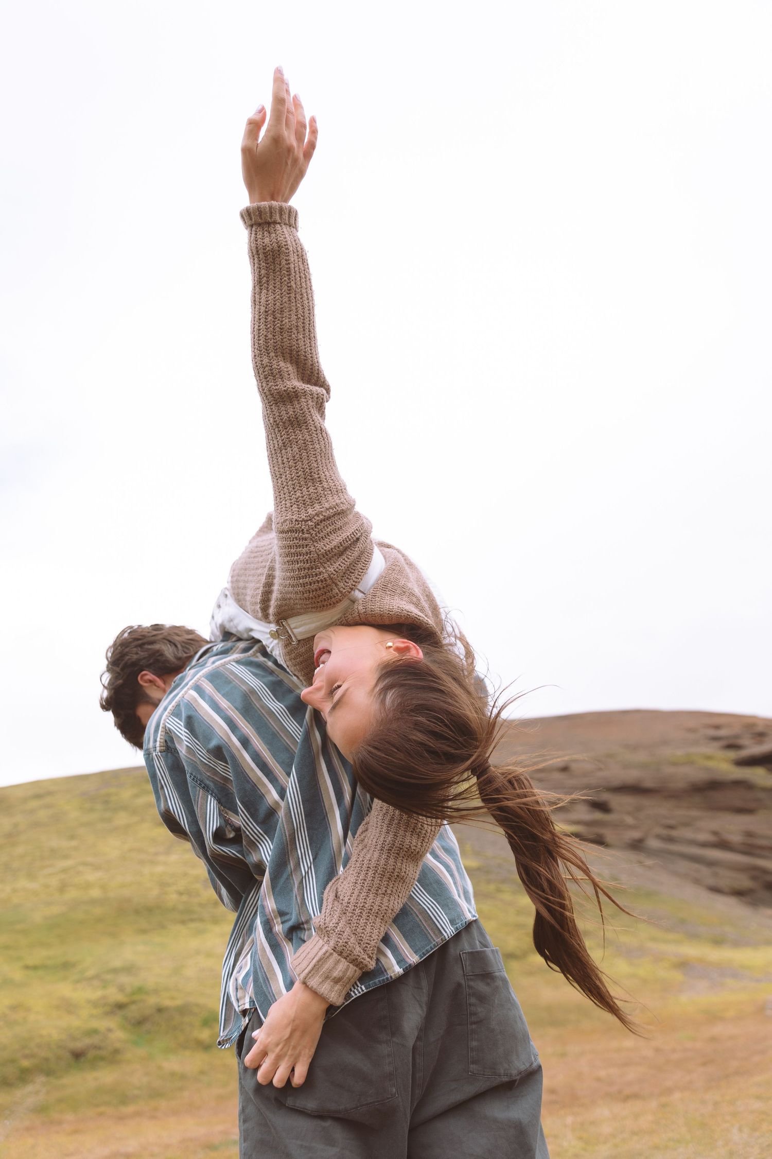 Woman reaching arm skyward while being carried on mans back in a dramatic open Icelandic hillside landscape.