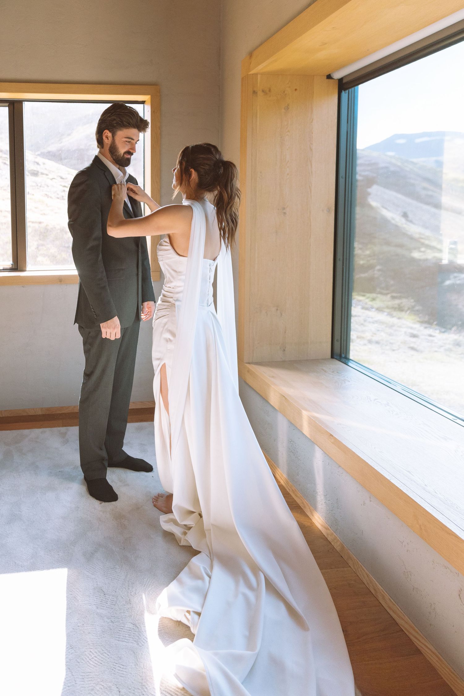 Bride and groom getting ready together by a sunlit window with mountain views.