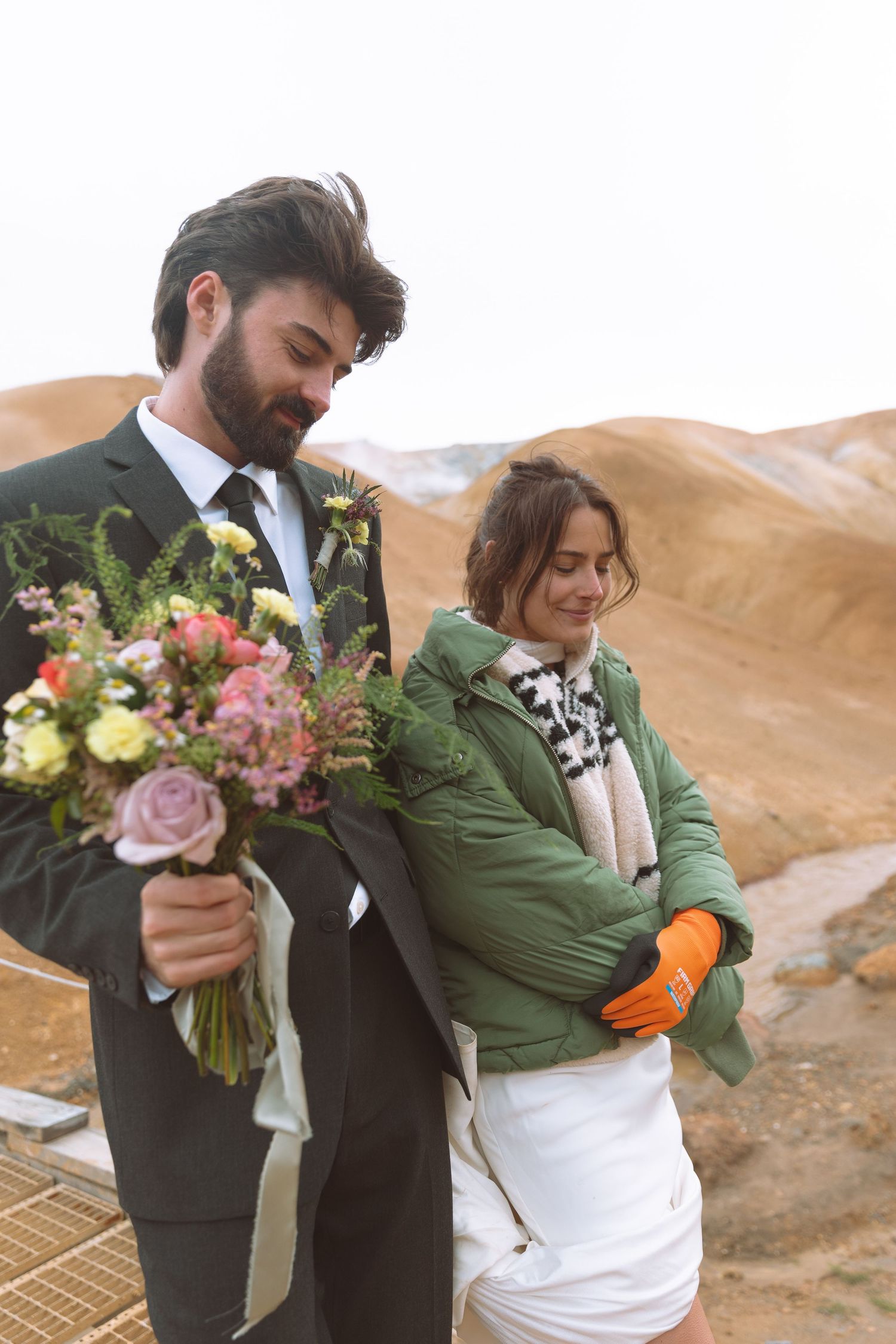 Groom in dark suit holds a colorful bouquet beside bride in white dress and green jacket in Iceland.