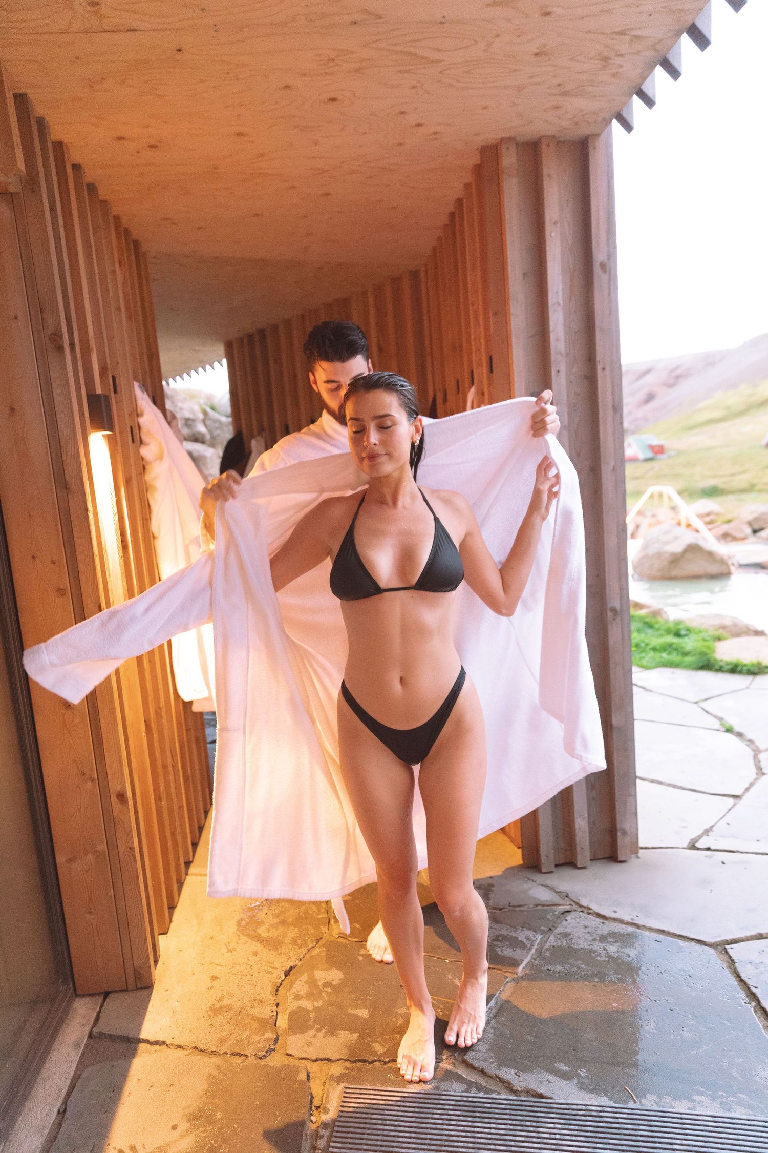 Woman in black bikini spreading white robe open in wooden spa corridor with man behind her.
