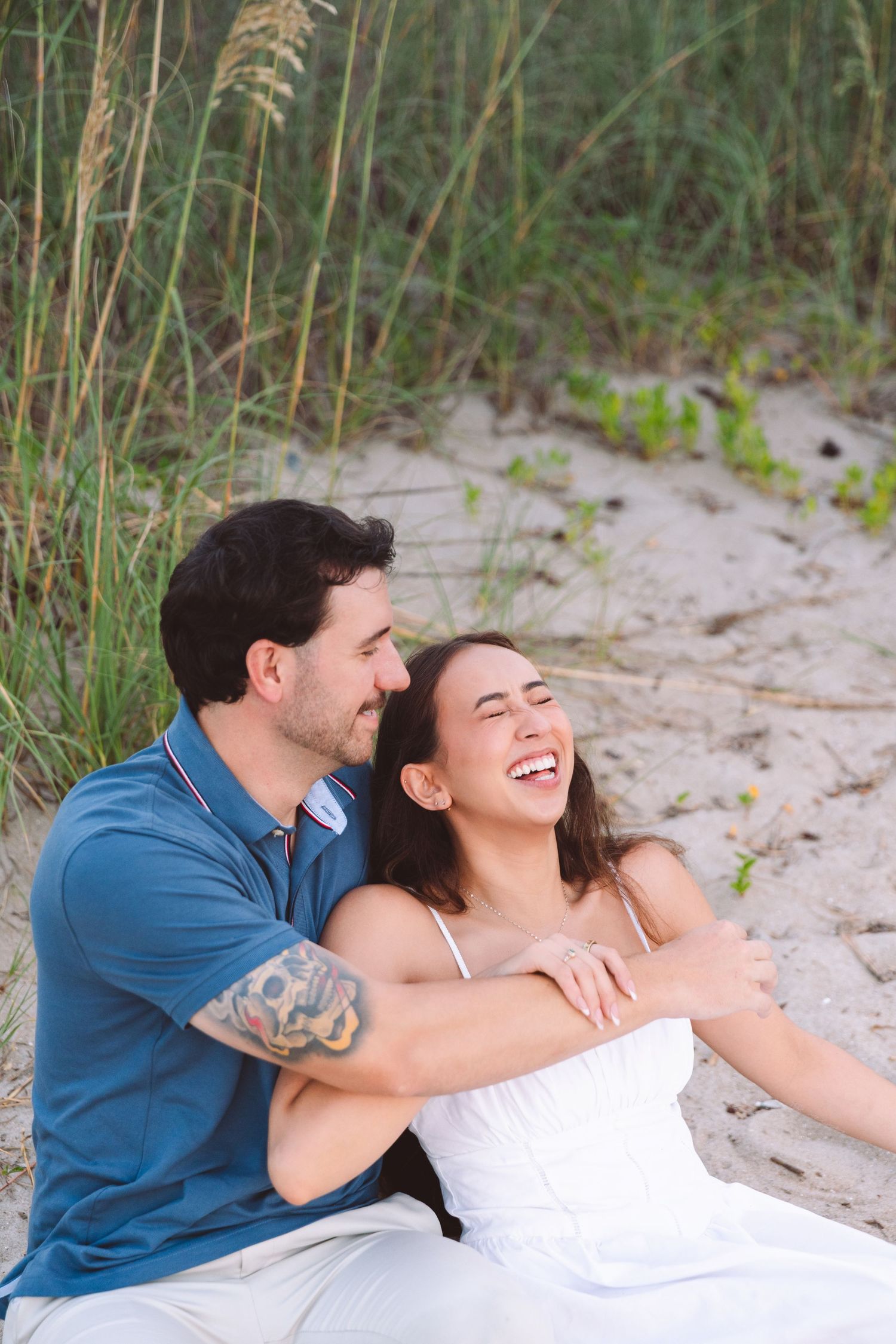 Happy couple laughing and embracing on sandy beach dunes surrounded by tall sea grass.