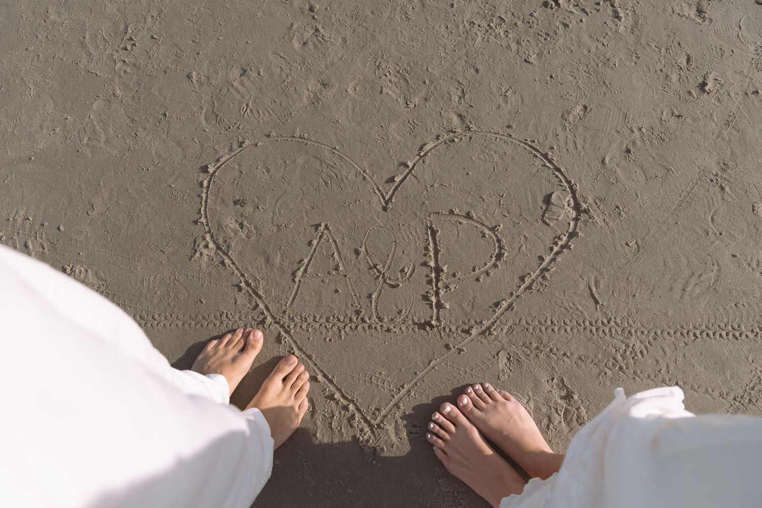Couple standing by a heart drawn in sand with initials at the beach.