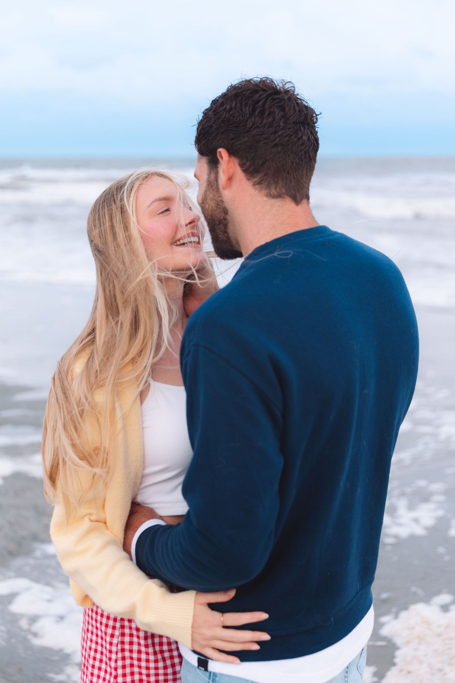 Smiling blonde woman embracing a man in a navy sweater on a windy beach.