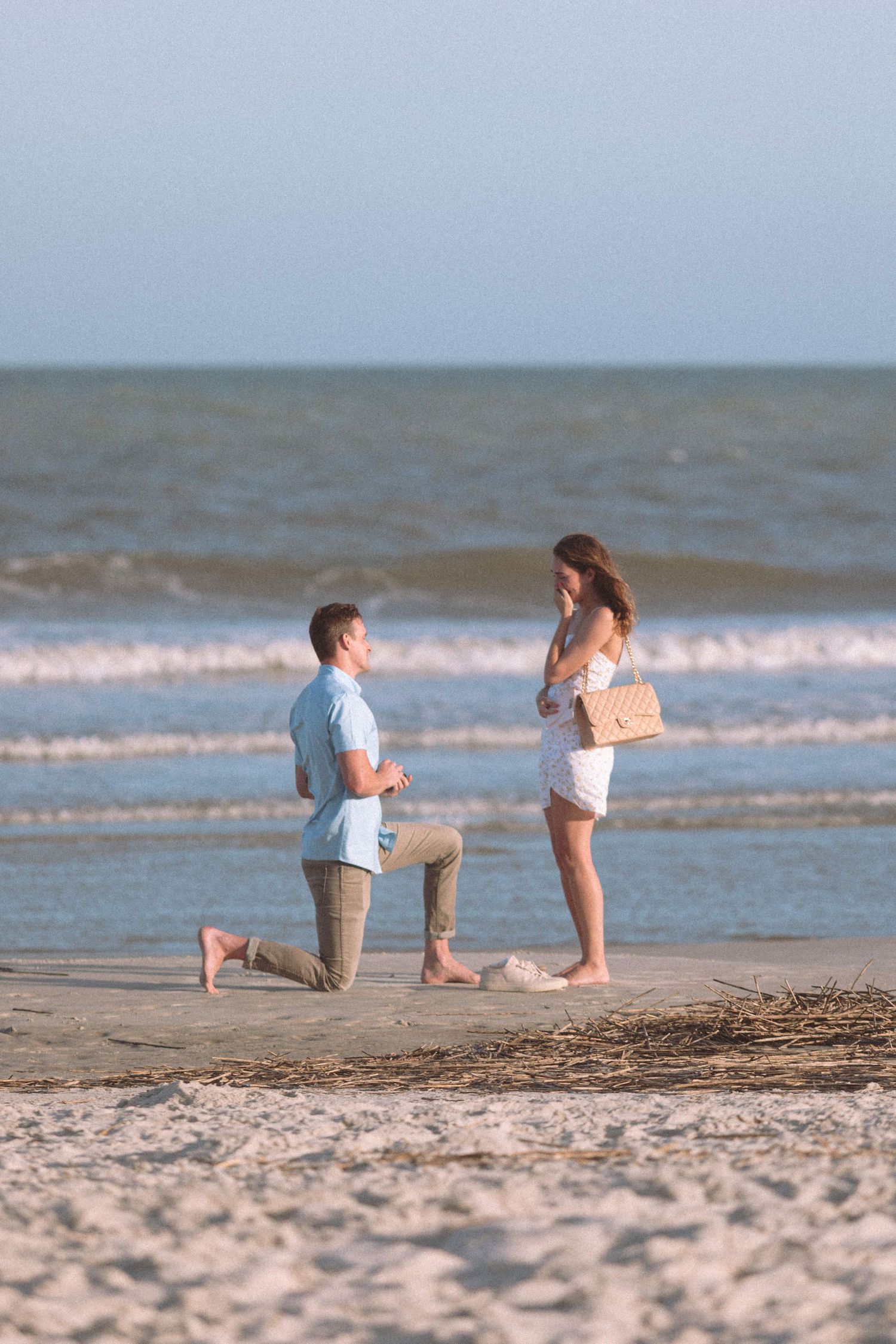 Man proposing on one knee to woman on a sandy beach at sunset with ocean waves in the background.