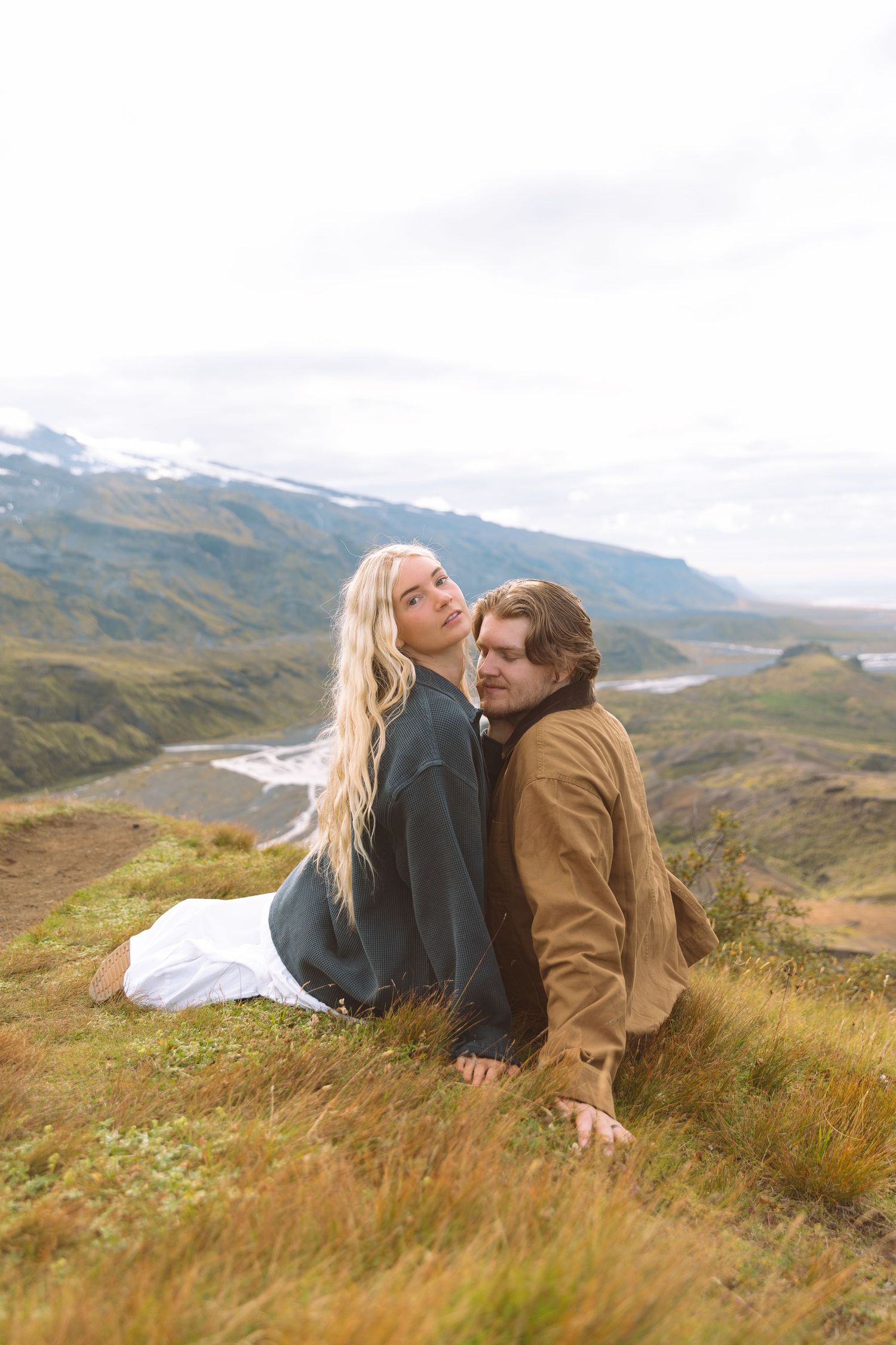 A blonde woman and a man sit closely together on a grassy hillside with a scenic mountain valley behind them.