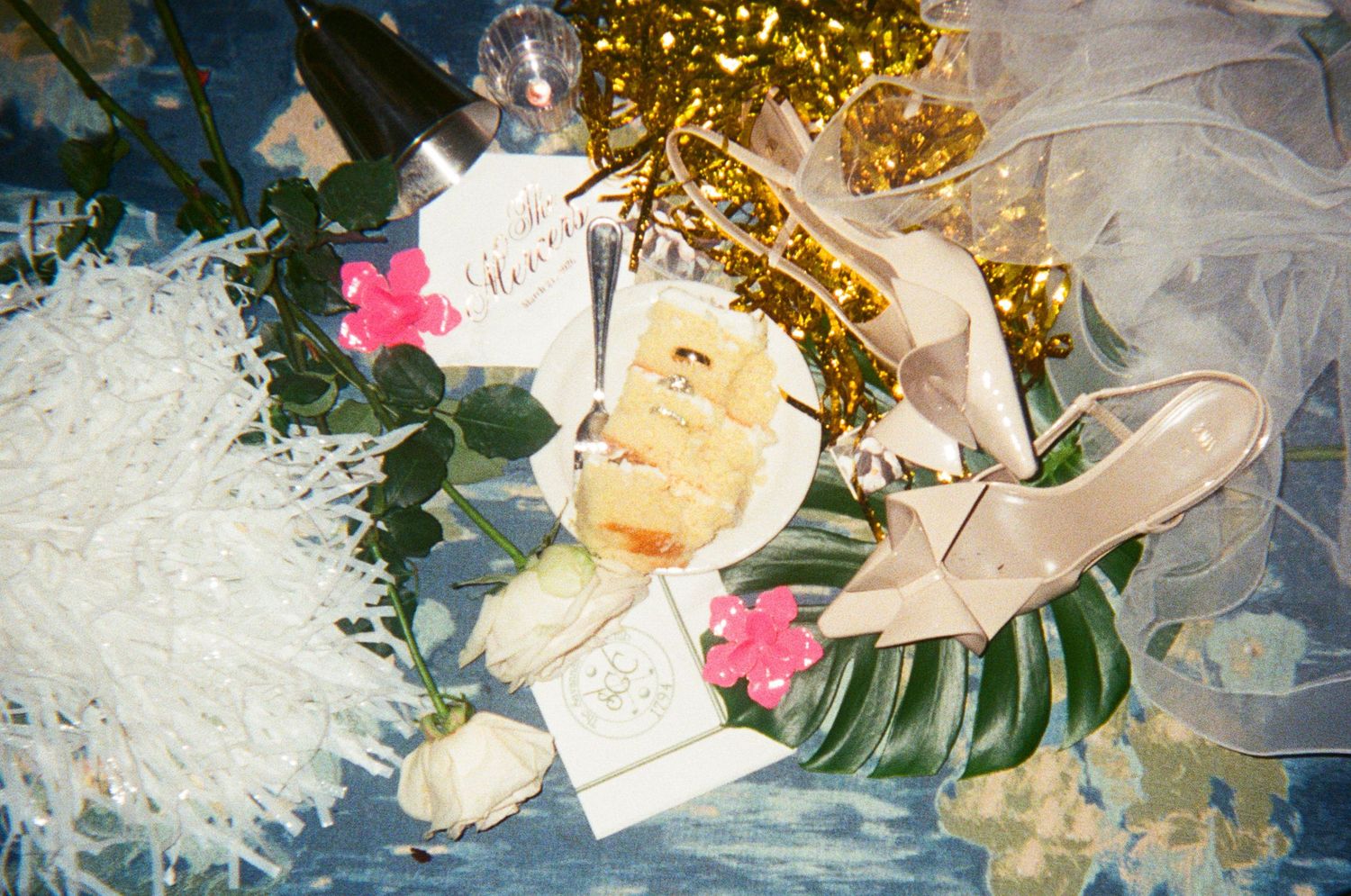 Flat lay of wedding details including white roses, pink flowers, heels, cards, pastries, and gold decorations on blue surface.