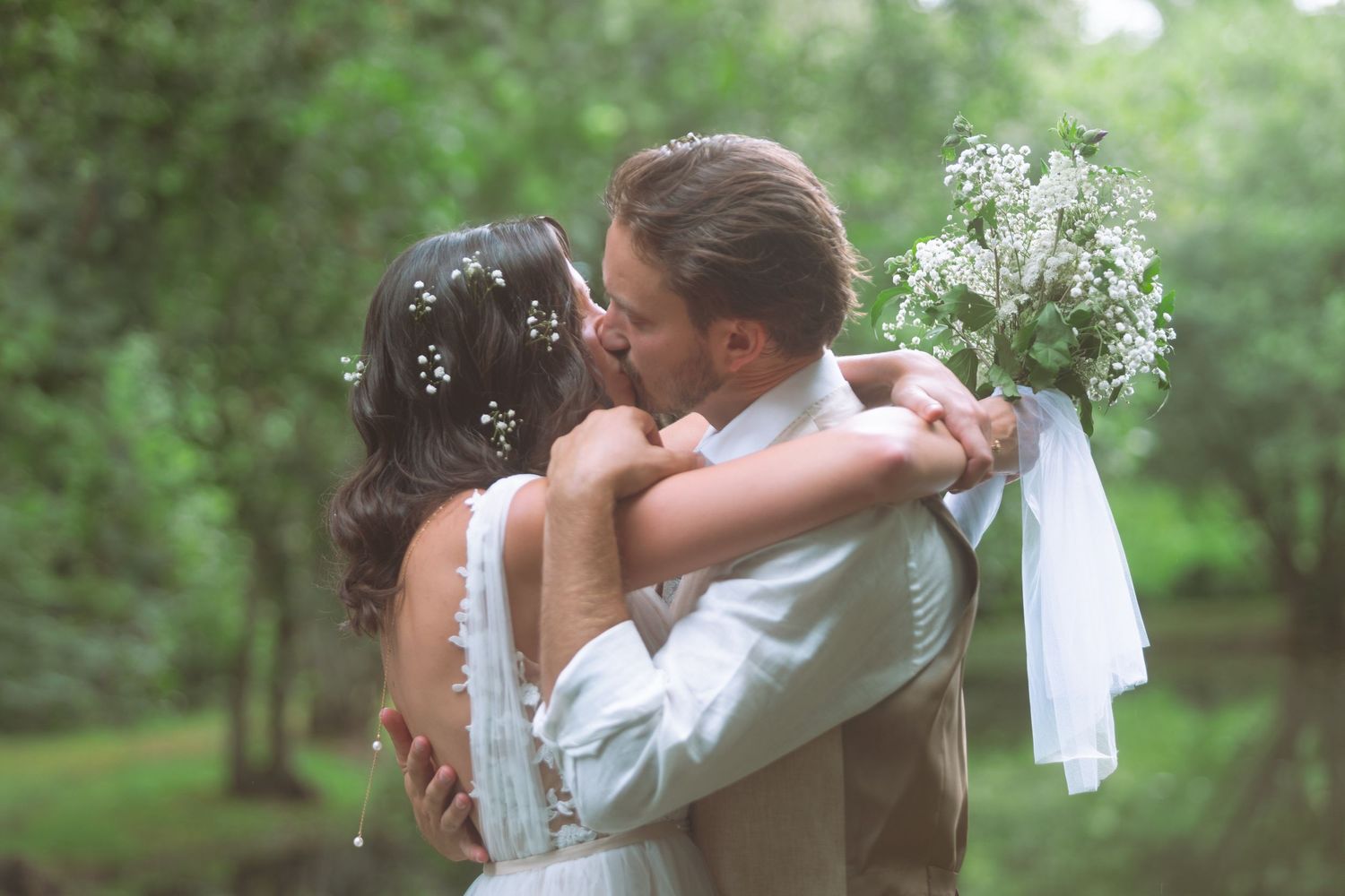 Bride and groom sharing a romantic kiss surrounded by lush green trees holding a white bouquet.