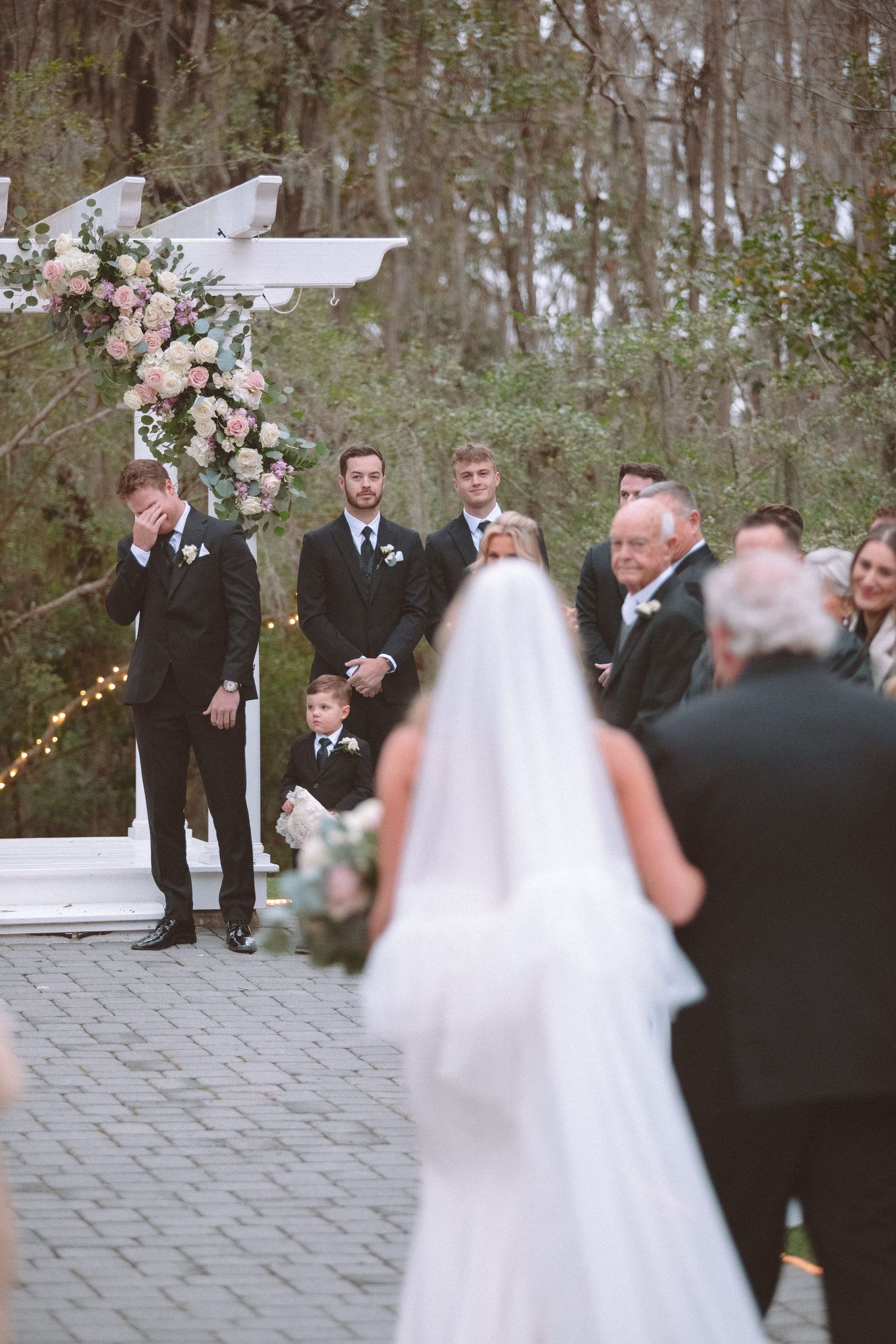 Emotional groom wipes tears seeing bride walk down aisle at outdoor woodland wedding ceremony.