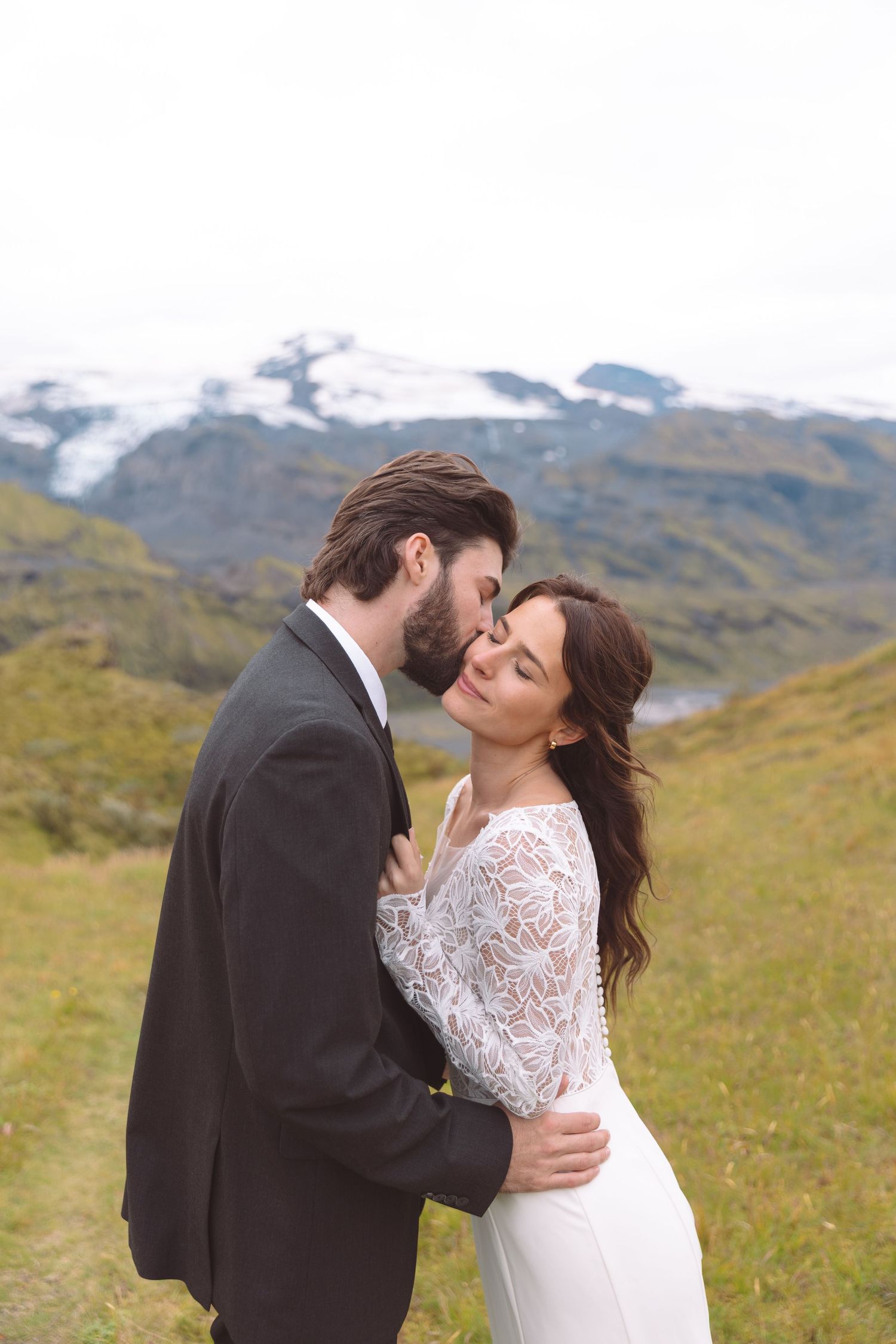 Groom tenderly kissing bride's cheek as she closes her eyes with stunning Icelandic glacier mountains behind.